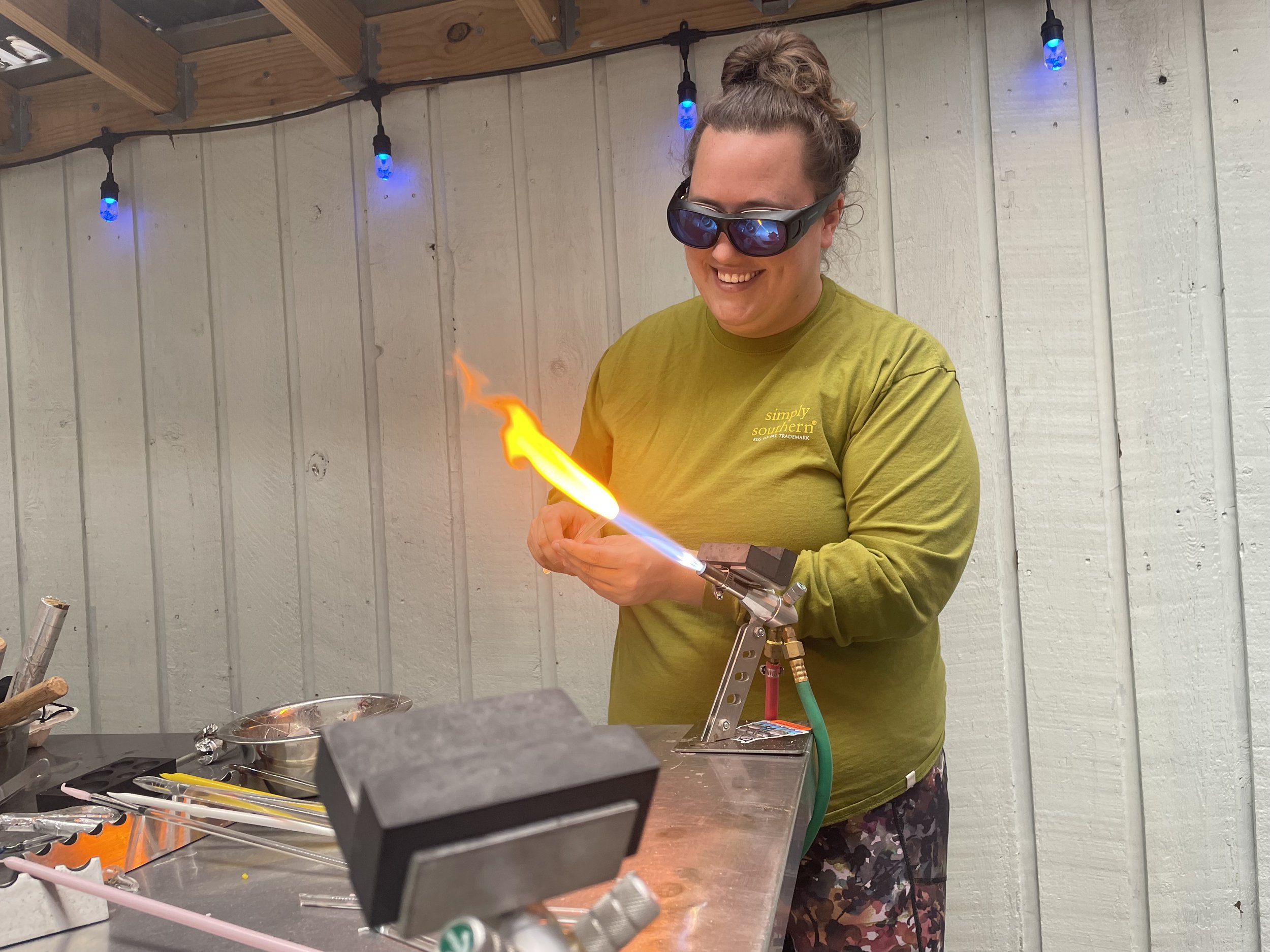 A woman in safety glasses and a green long-sleeve shirt is holding a flame torch, which is creating a visible flame, in a workshop or studio with wooden ceiling beams and string lights. She is smiling and appears to be engaged in a glassblowing or me