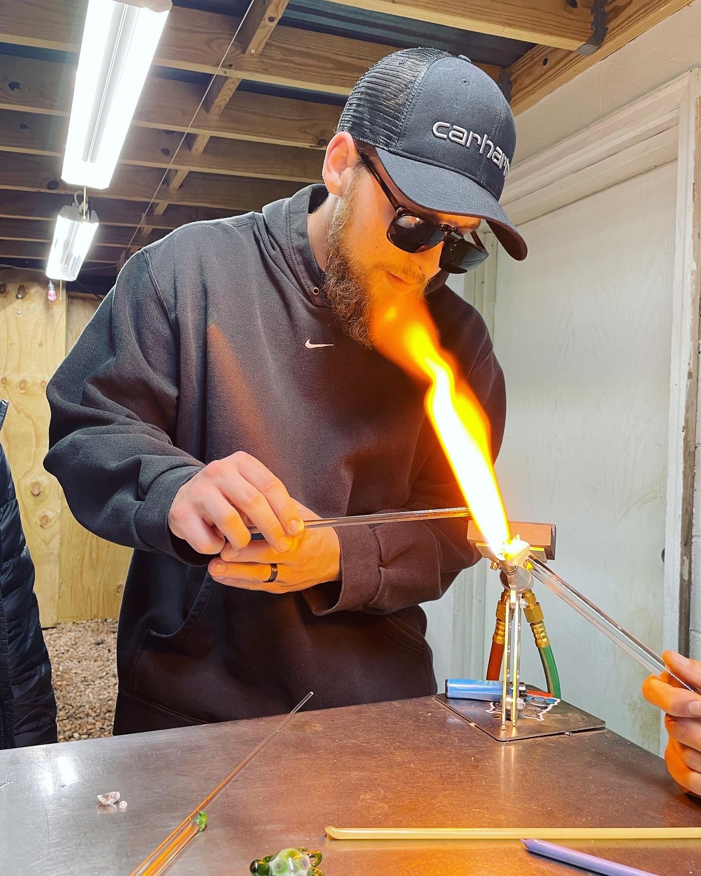 A man wearing a black hoodie, sunglasses, and a hat, using a torch to work on a glassblowing process, creating a flame inside a workshop.