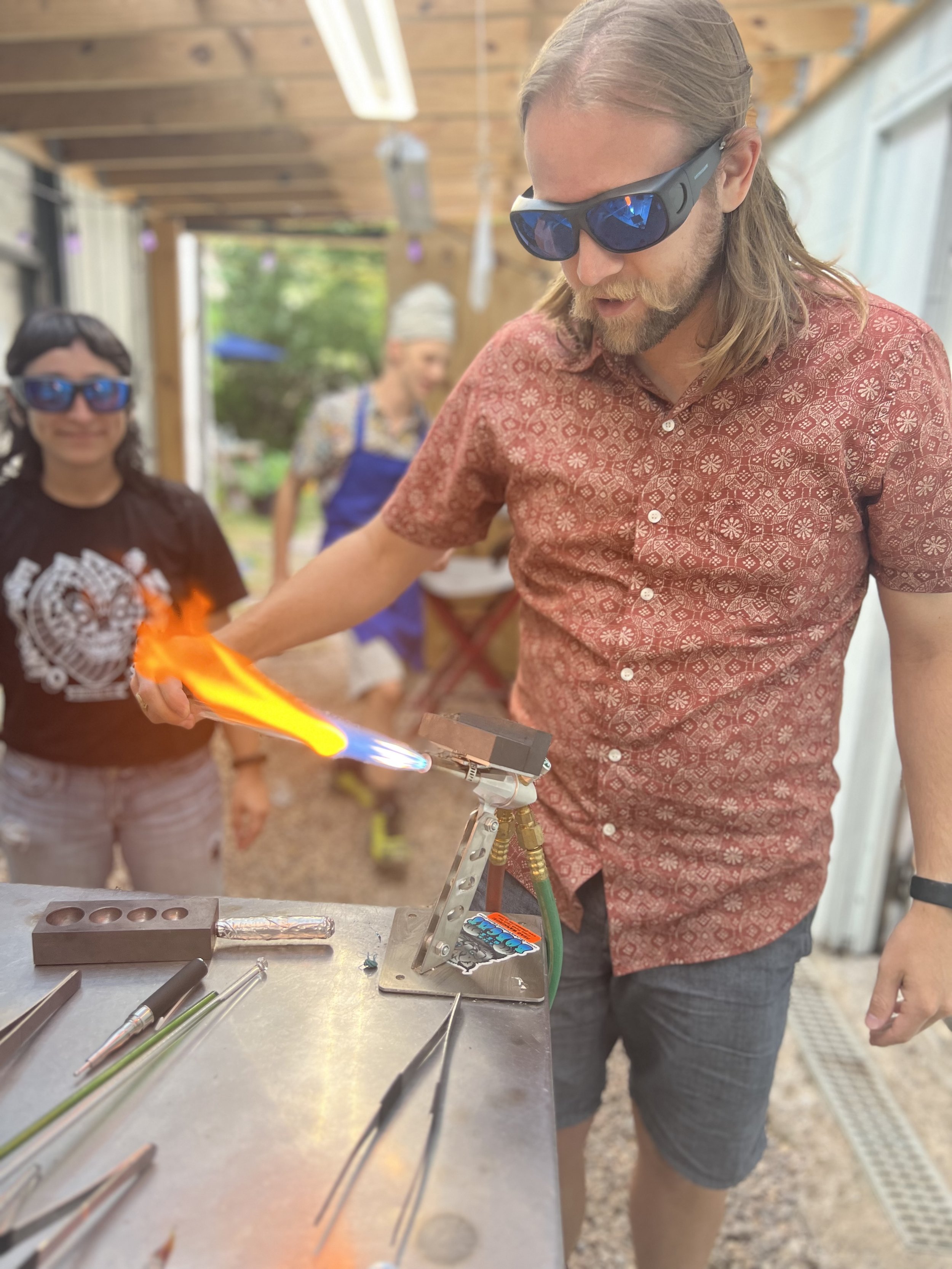 A man wearing safety glasses and a patterned red shirt is using a blowtorch to work on glass art, with two women in the background observing, in a workshop or outdoor workspace.