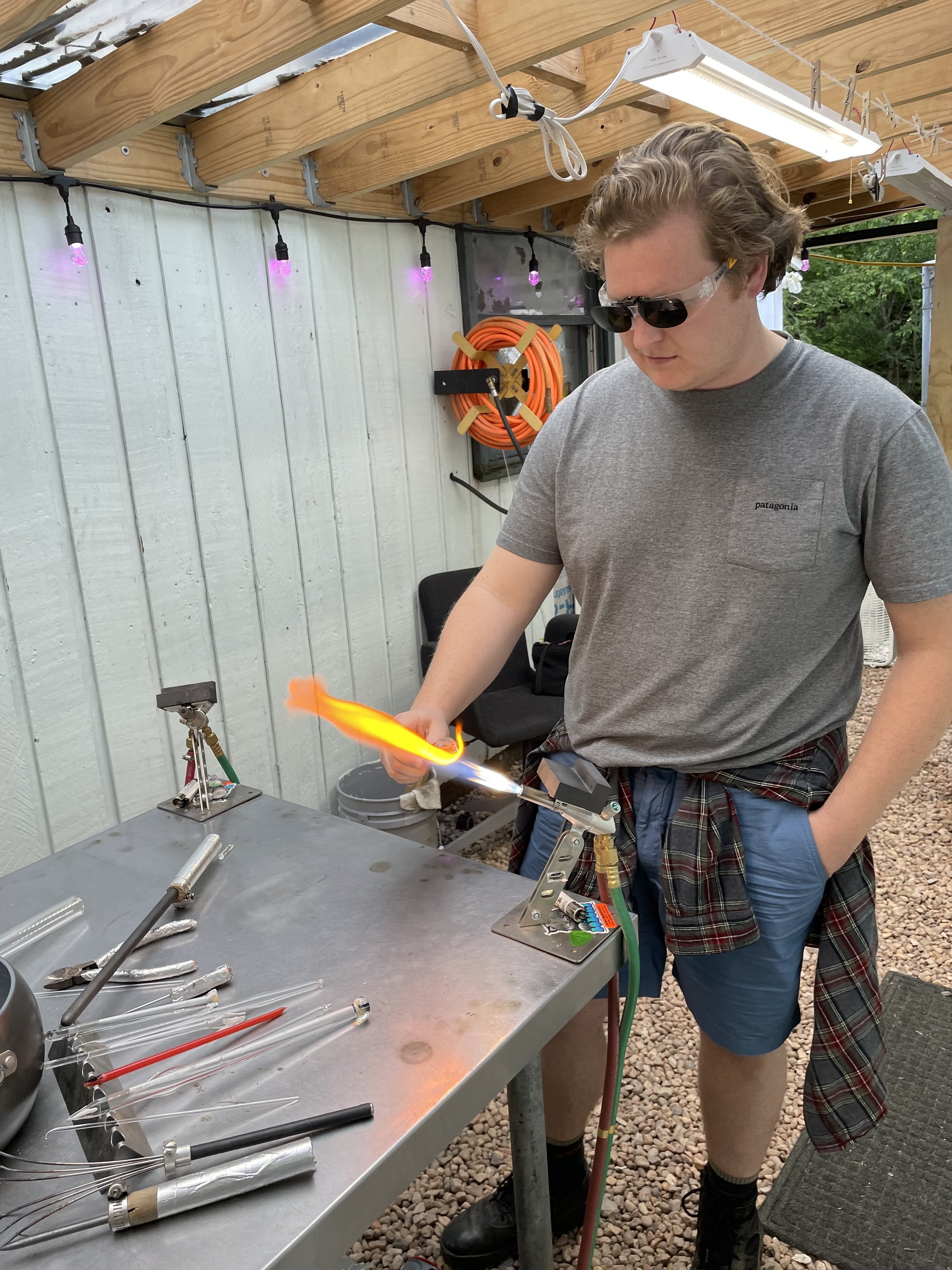 Man wearing sunglasses and casual gray t-shirt working on glassblowing, using a torch at a metal workbench with glass tools, in a workshop with string lights and outdoor setting.