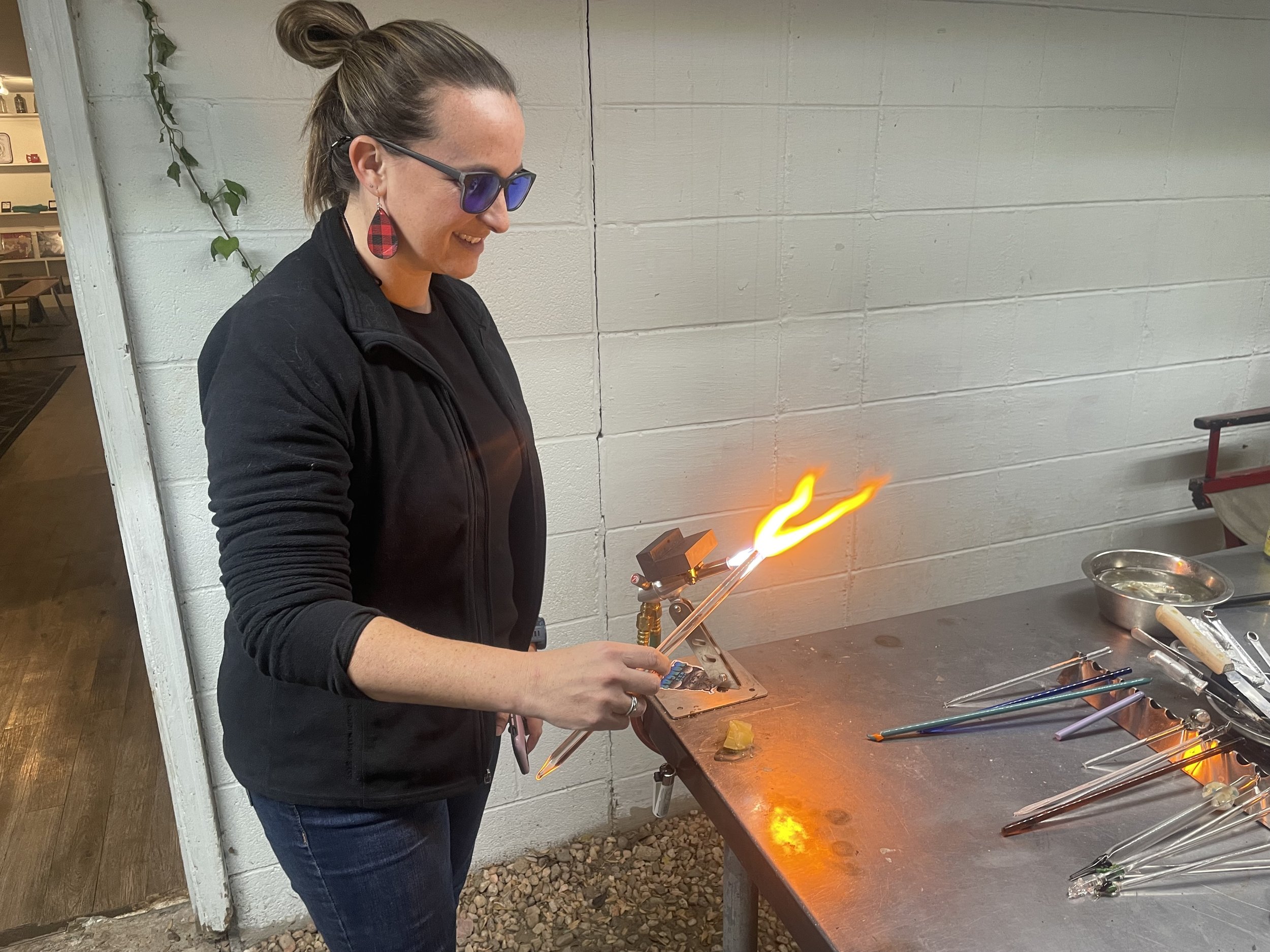 A woman wearing glasses and a black jacket is working with glass in a glassblowing workshop. She is holding a long glass tube that is emitting a flame, shaping it in the air. Various tools and glass pieces are on the metal worktable.