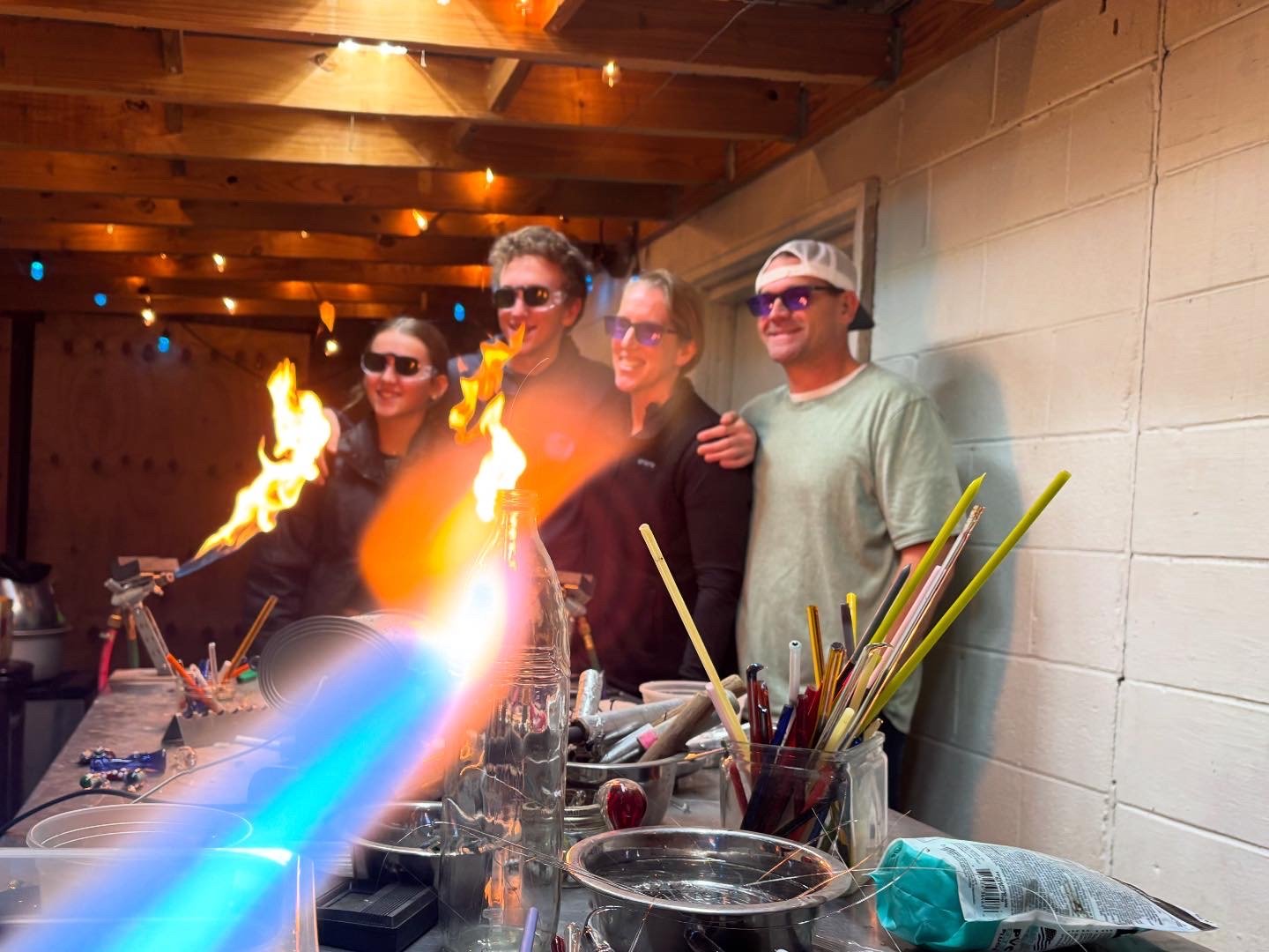 Four people wearing sunglasses smiling in a workshop decorated with string lights, with a table holding glassware and tools in front of them. Flames are coming from a bottle on the table, indicating a science experiment or demonstration.