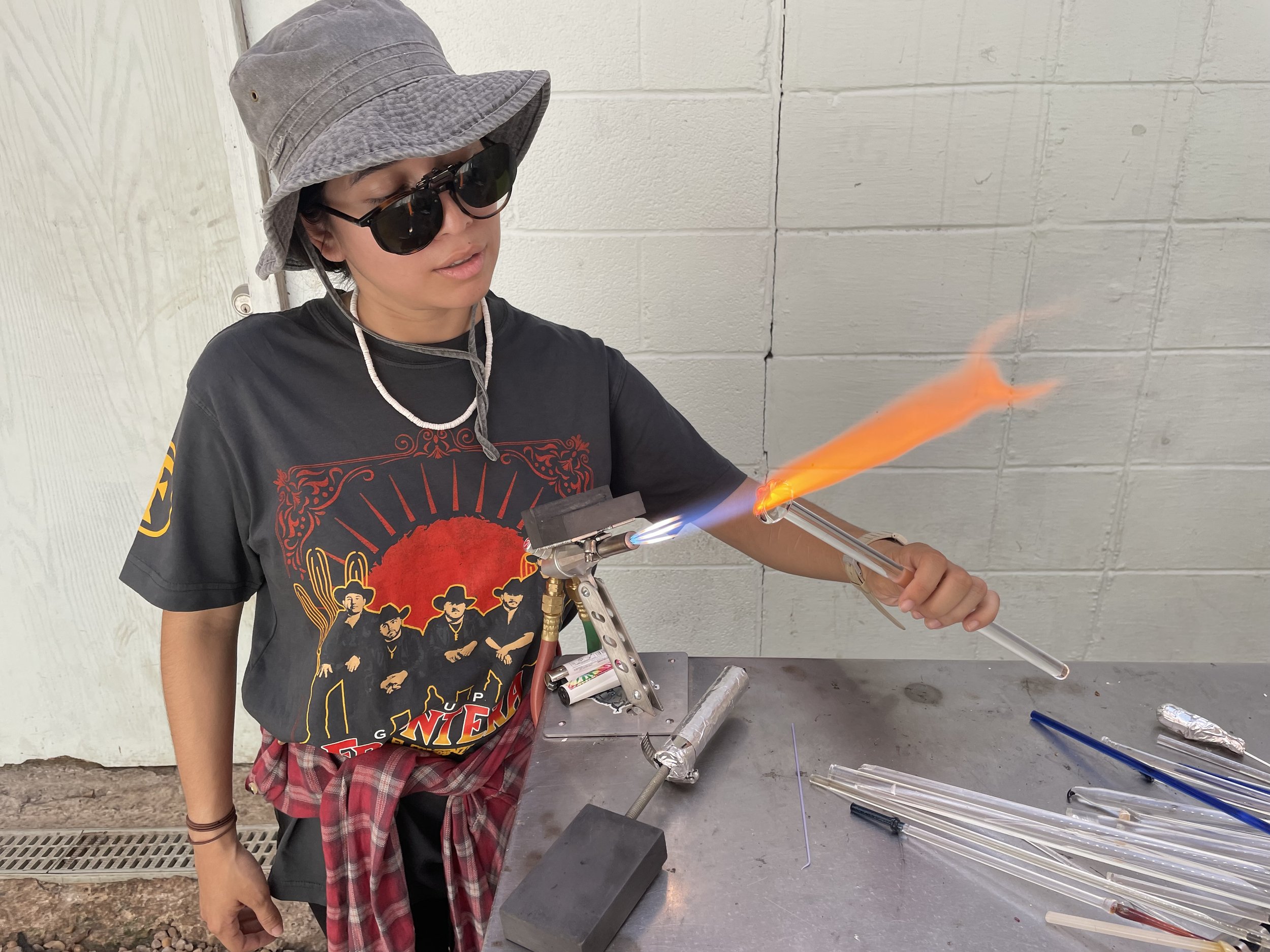A young woman wearing sunglasses, a gray sun hat, and a black T-shirt with a colorful graphic design, is holding a glass tube with a flame at one end, performing glassblowing or glassworking inside a workshop.