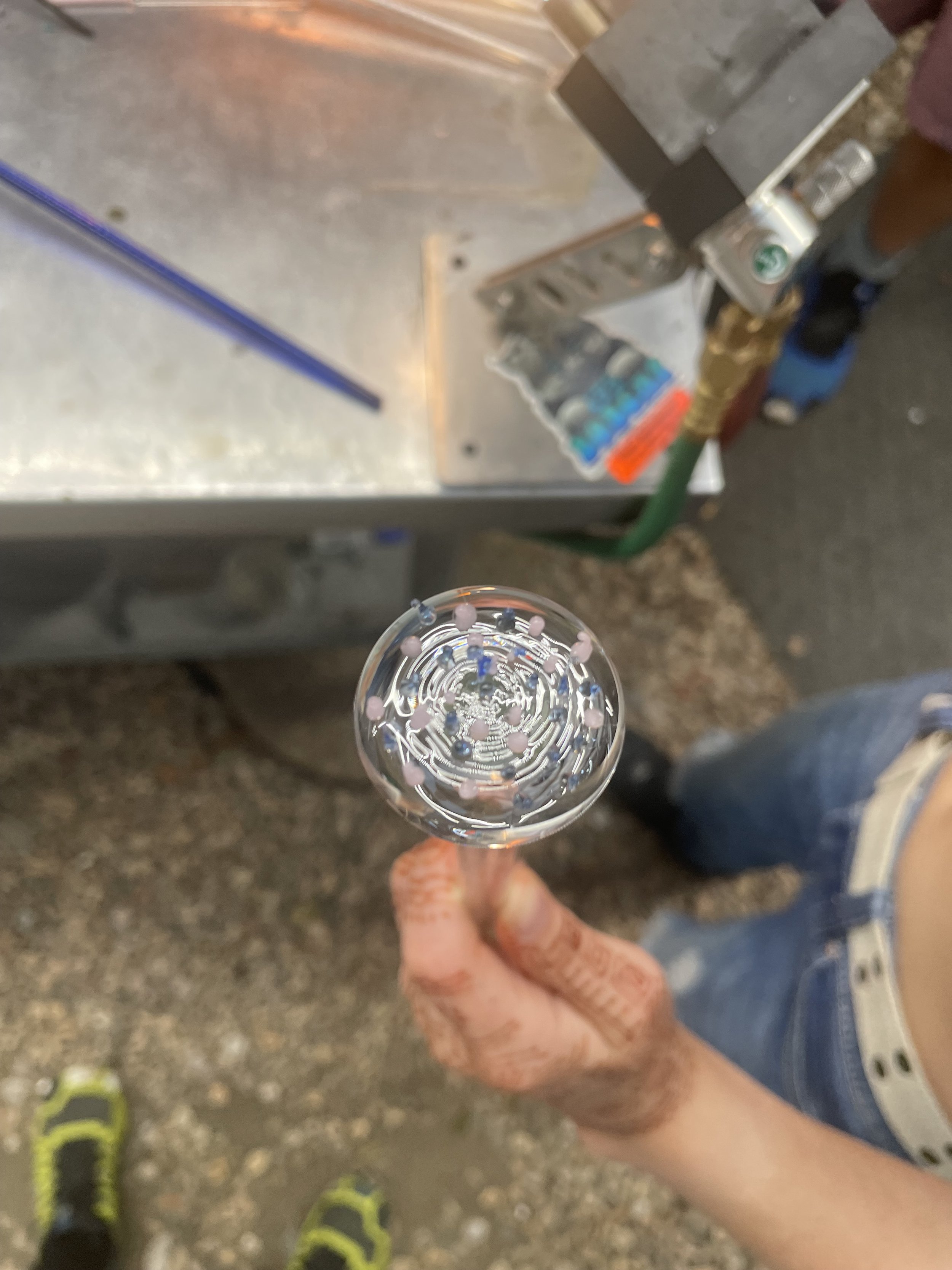 A person holding a glass of clear liquid with small colorful beads inside, with a metal workbench and tools in the background.