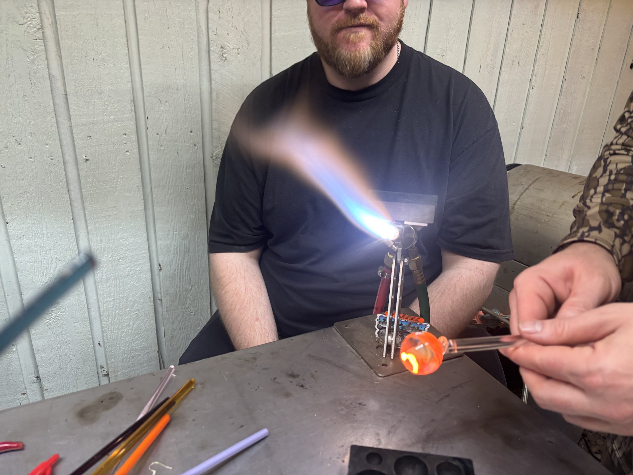 A man with a beard wearing a black T-shirt sits at a metal workbench while another person is holding a glass tube with an orange glow near a small flame or torch, engaged in glassblowing or lampworking, with colorful glass rods on the table.