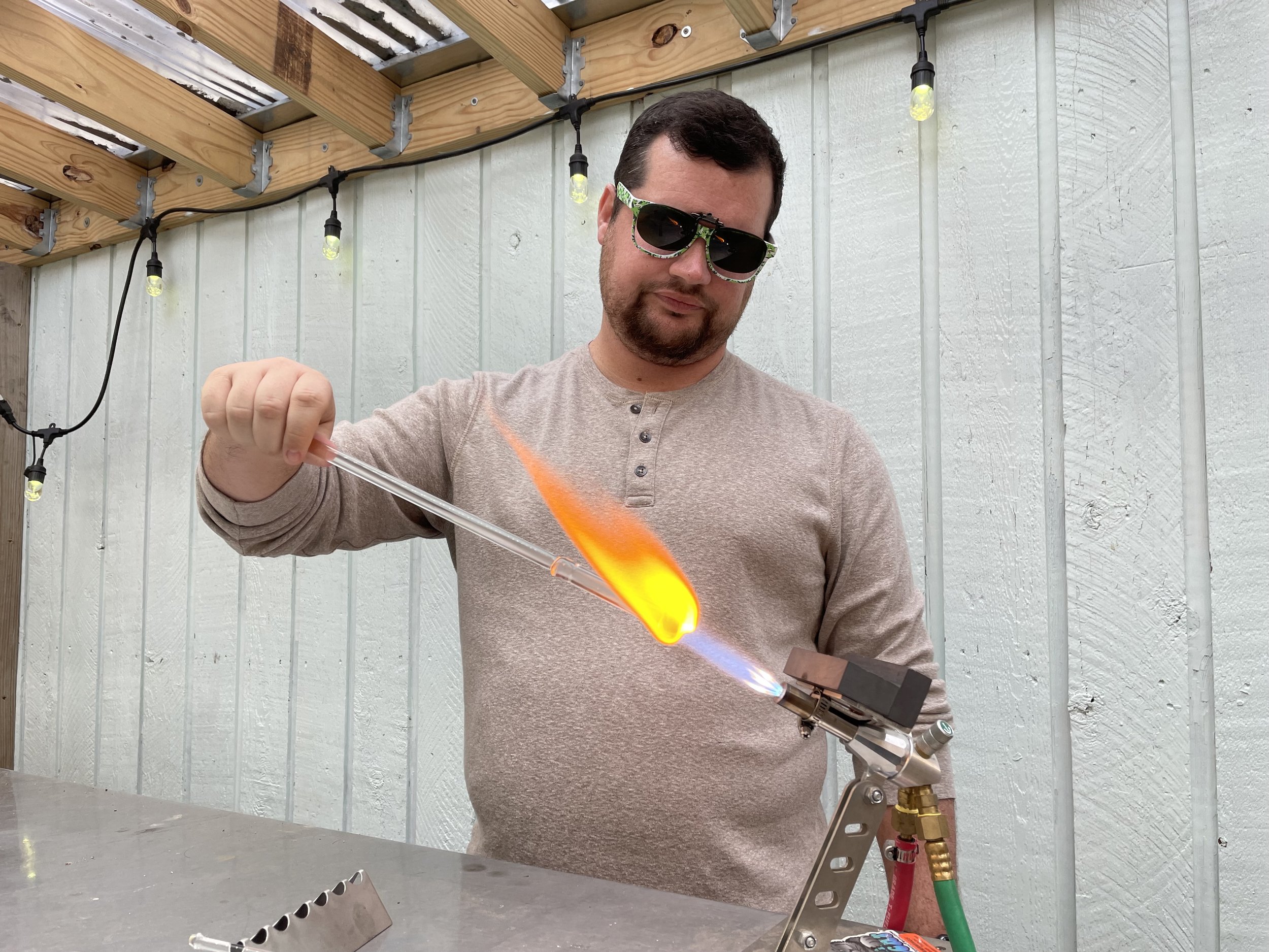 A man wearing sunglasses and a beige long-sleeve shirt is holding a glass tube in a flame produced by a propane torch, with a clothespin holding the tube. He is working outdoors near a light-colored wooden wall with string lights overhead.