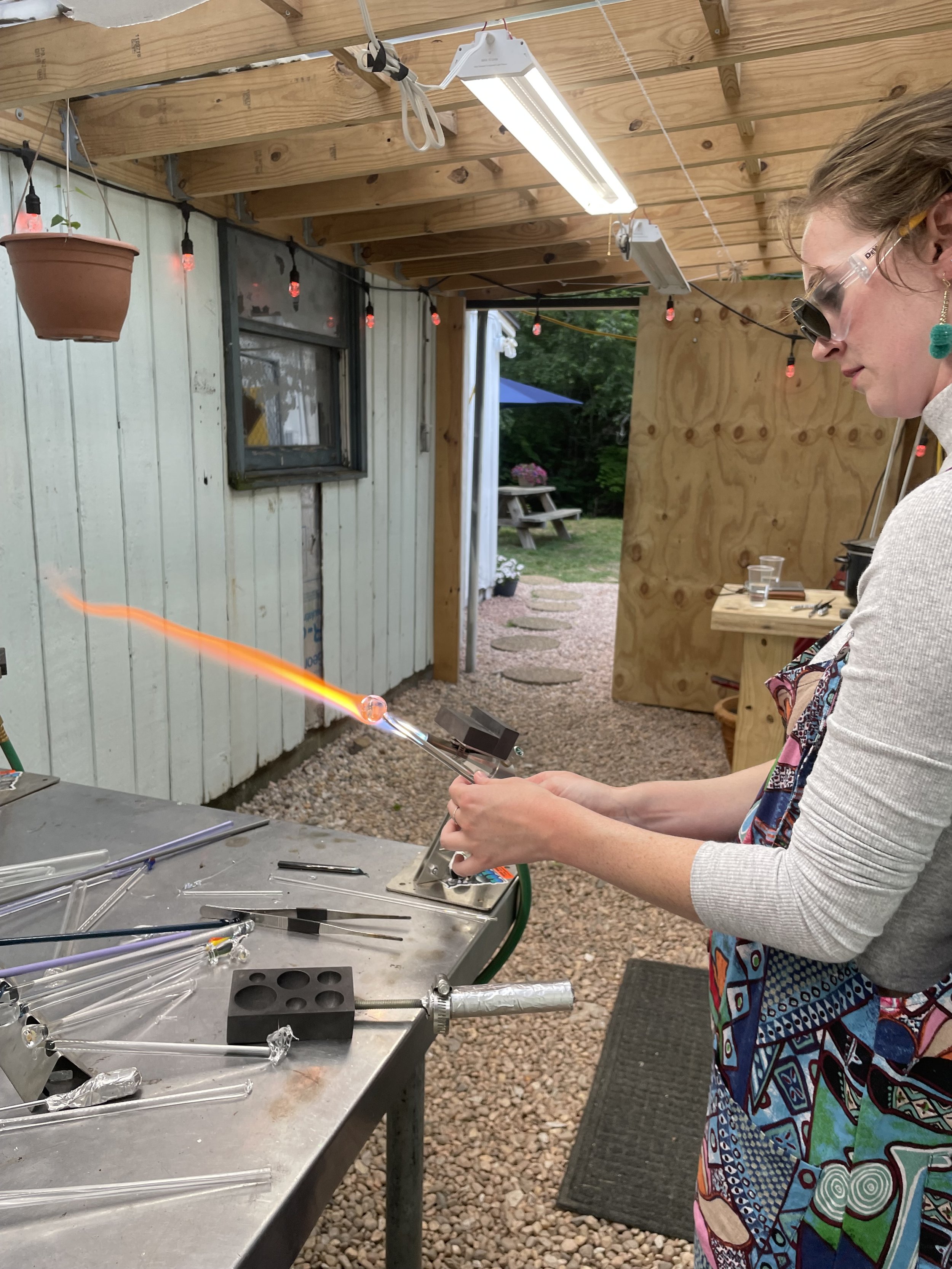 A woman wearing safety glasses and a colorful apron is working with glassblowing tools to shape molten glass in a workshop. There are various glass rods and tools on the metal table.