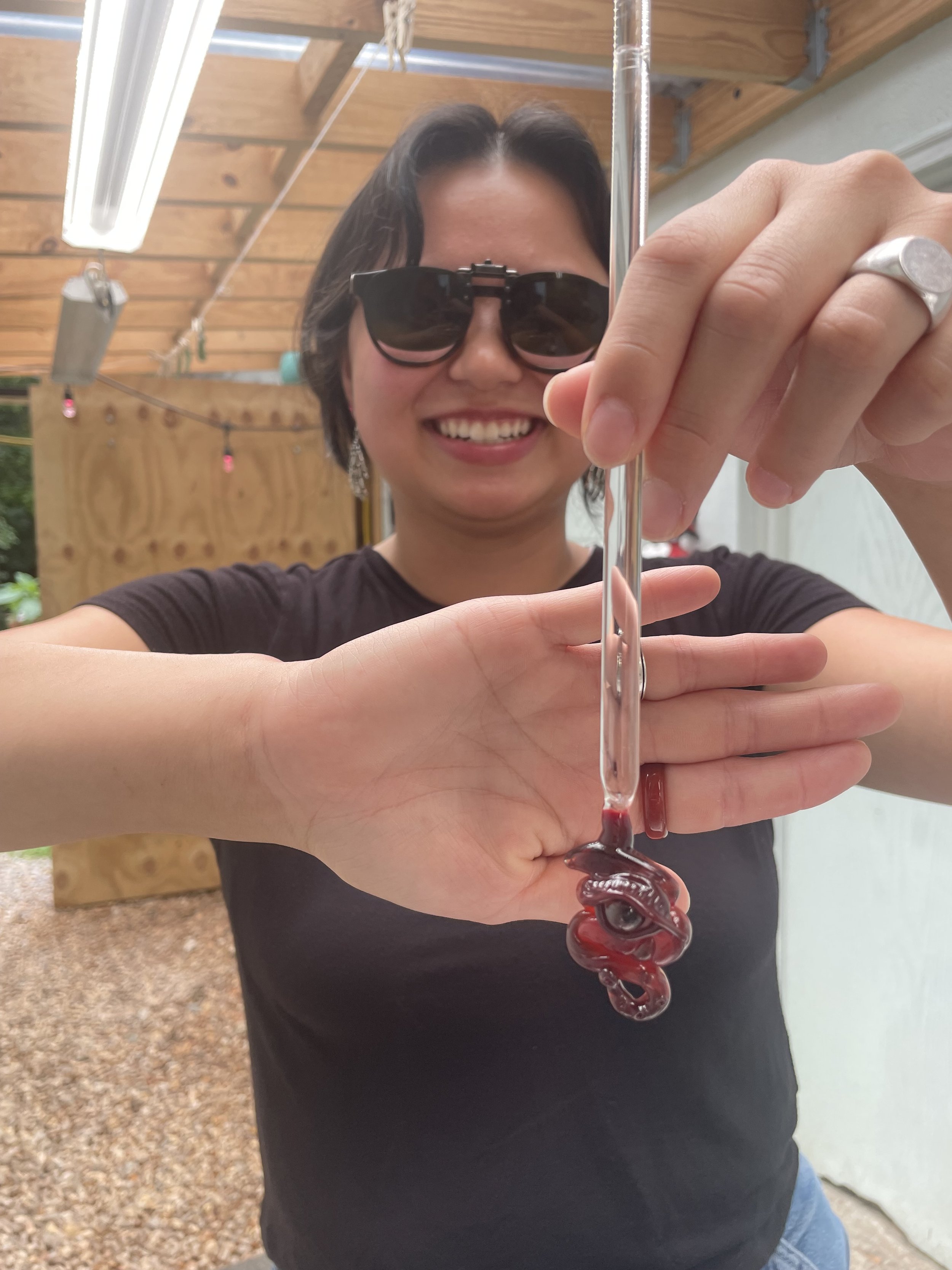 Woman smiling, wearing sunglasses, holding a glass pipette with a red liquid inside, in an outdoor setting with a wooden wall and string lights.