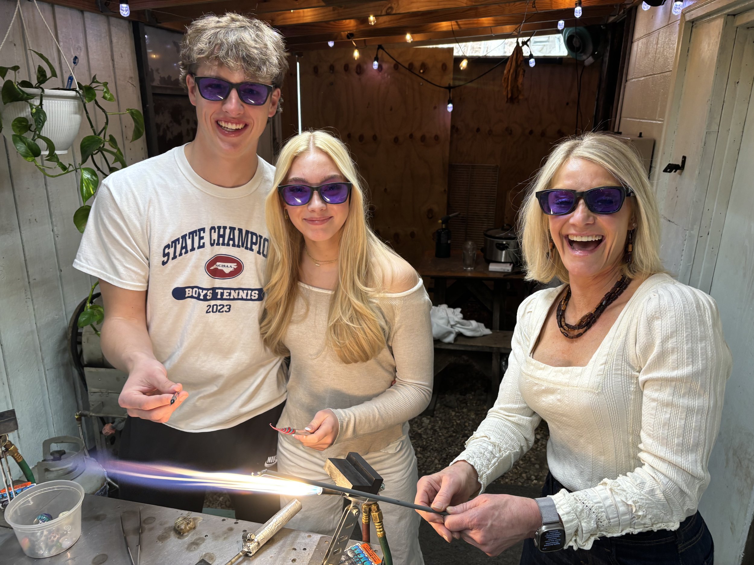 Three people in a workshop, wearing purple safety glasses, are smiling while candle making. The woman on the right is holding a lit candle wick over a torch, and the other two are standing nearby.