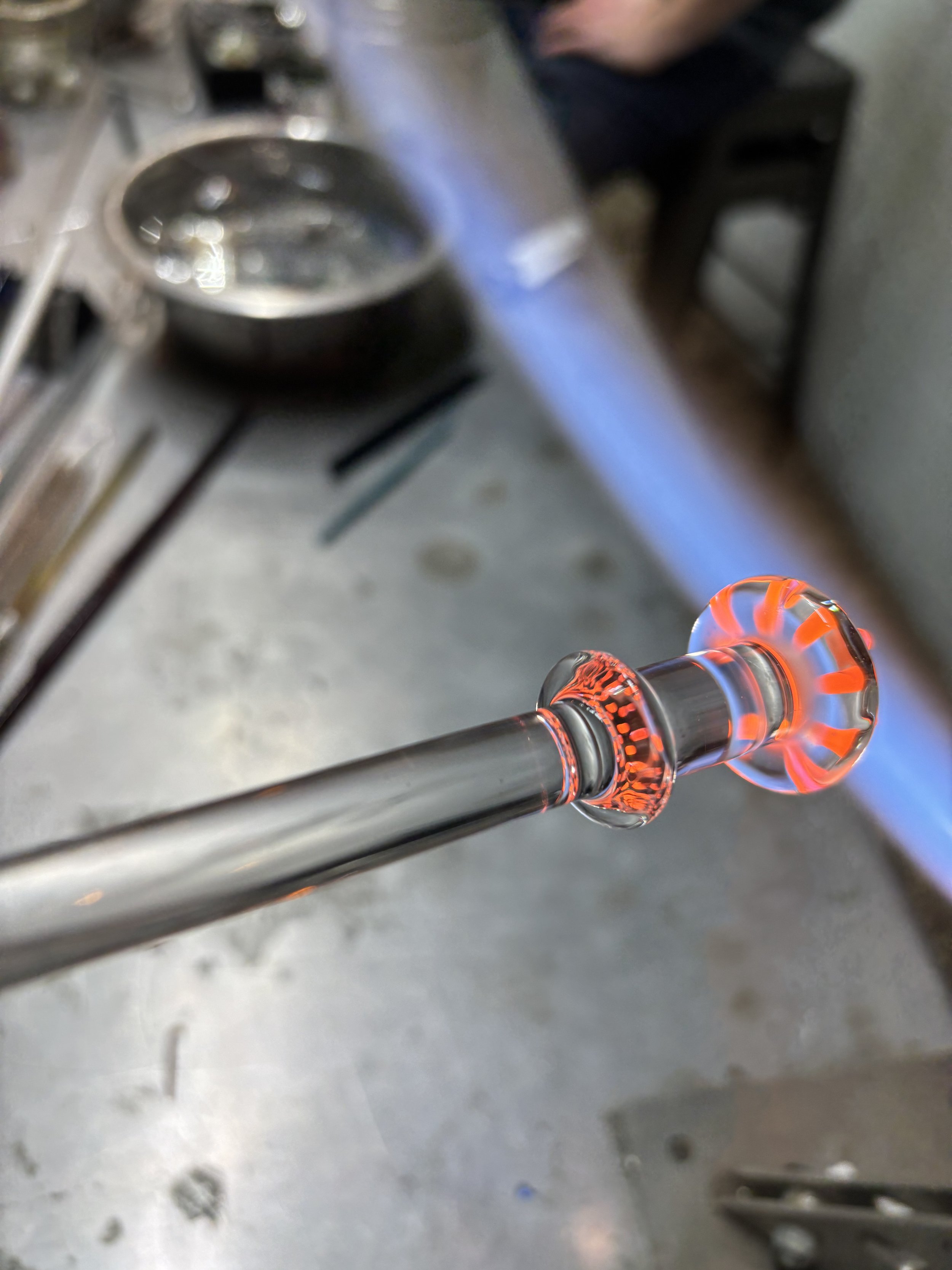 A glassblower is shaping a piece of molten glass into a decorative object in a workshop. The glowing orange glass is attached to a metal blowpipe.