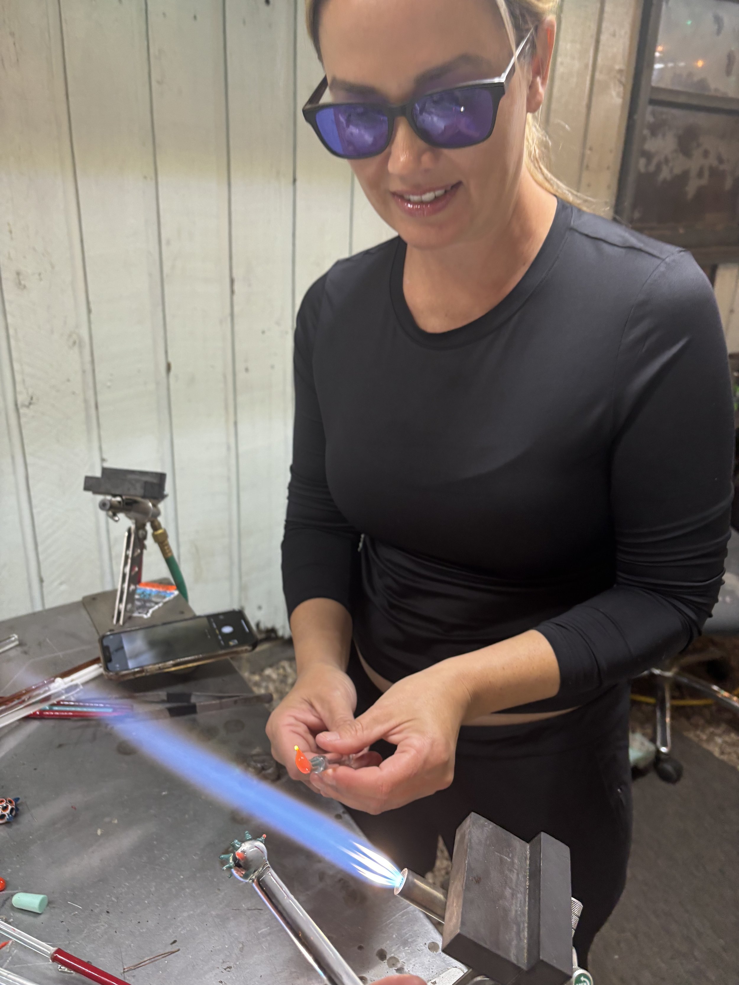 Woman working with glassblowing torch, wearing protective glasses, shaping glowing glass at a workshop.