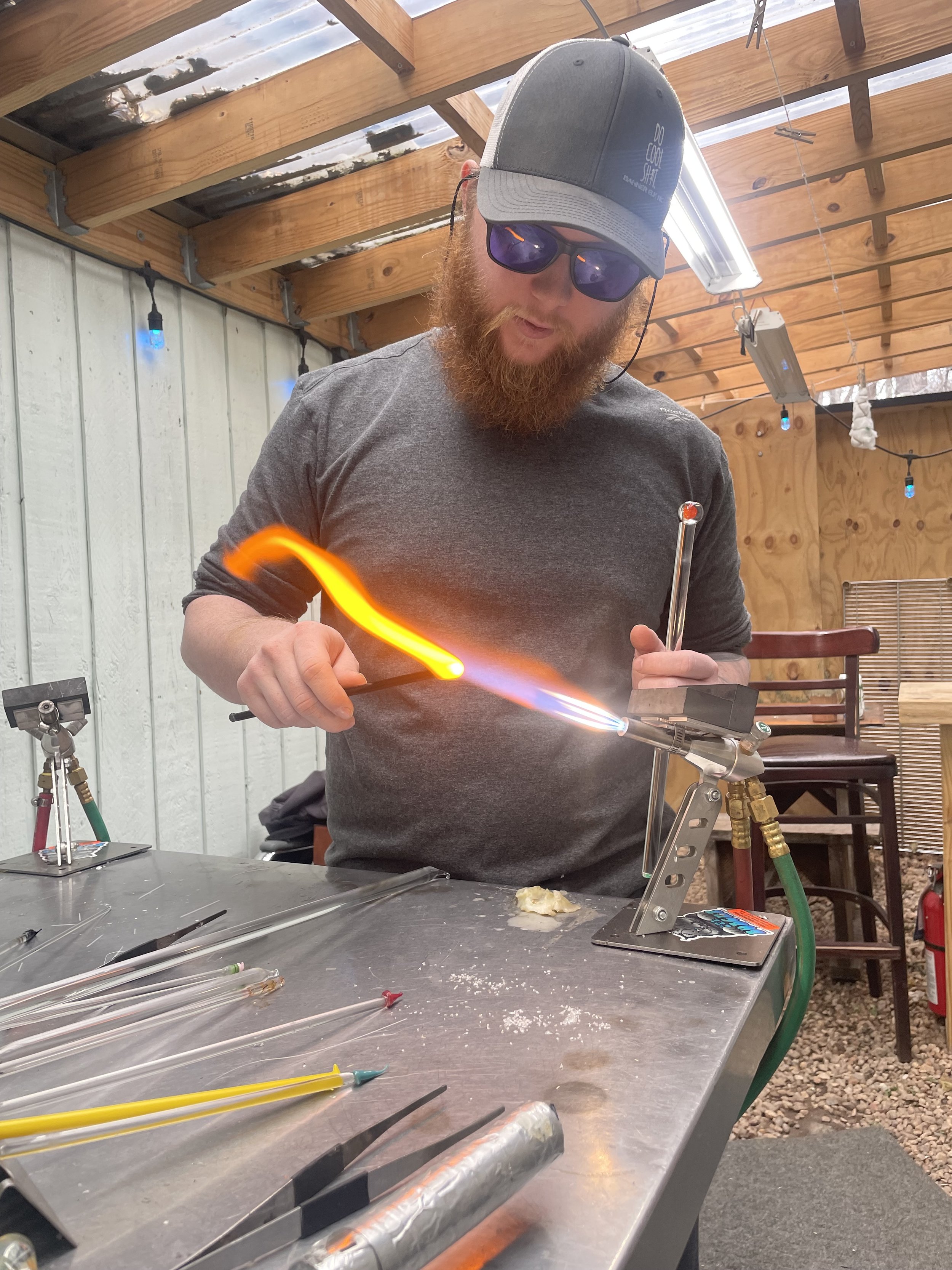 A man with a red beard wearing sunglasses, a cap, and gray shirt is working on glassblowing at a workbench. He is holding a glass rod in his left hand and a tool in his right hand, shaping glowing orange glass with a torch. Various glassblowing tools