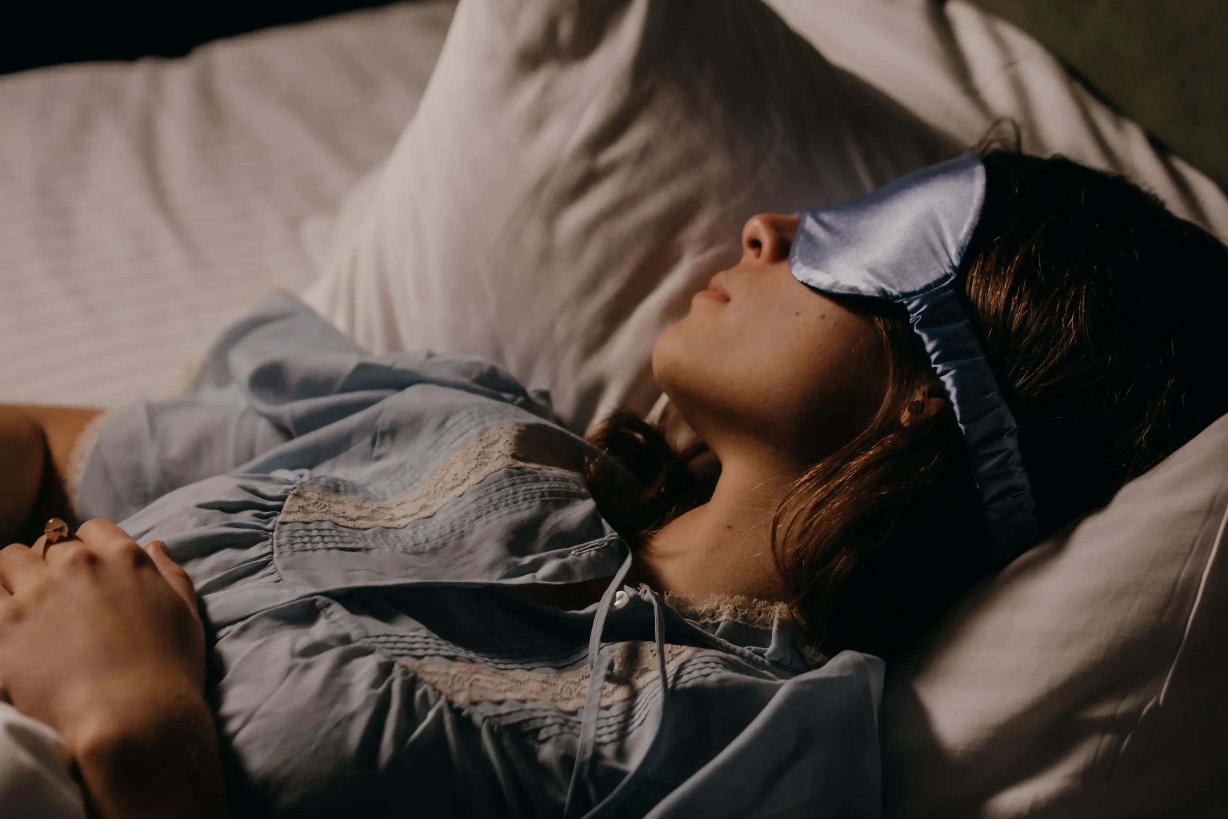 Woman lying in bed wearing a blue satin sleep mask, with her hands resting over her abdomen.