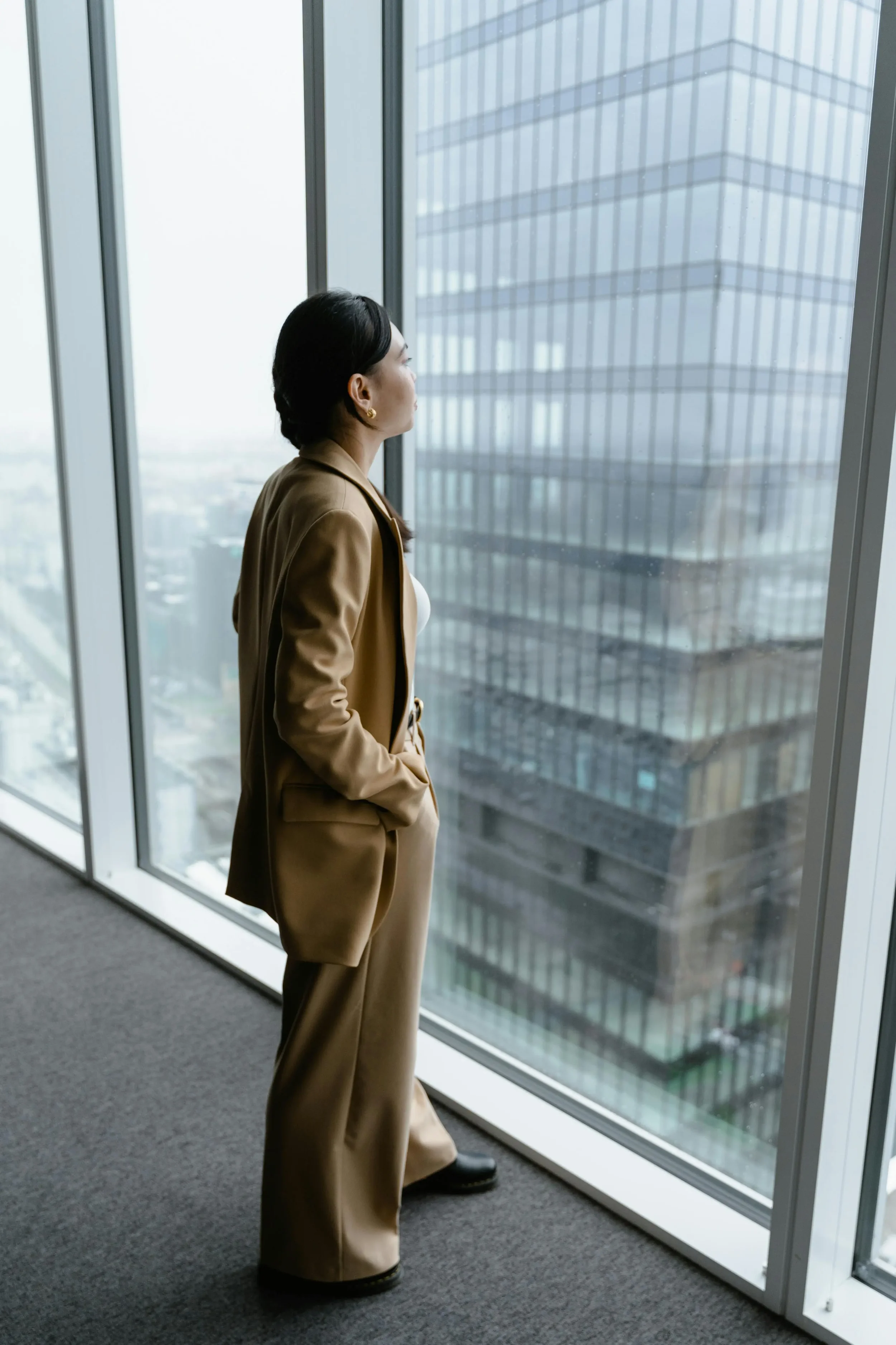 Professional woman looking out a large office window at city buildings, suggesting reflection, stress or career pressure