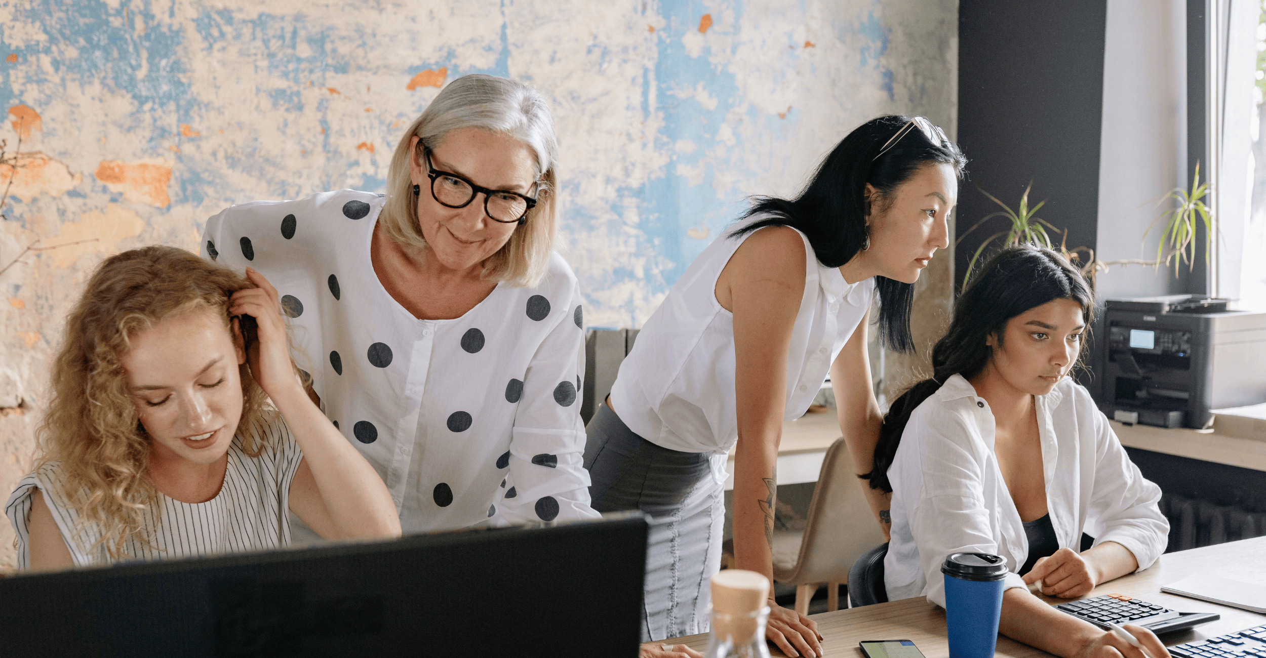 Four women in the office, two each working on laptops, two older ladies standing up behind, looking over their colleagues shoulders, smiling.