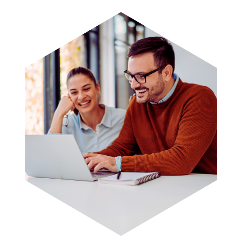 Two office workers- one male and one female, sitting working at a shared laptop, both are smiling.