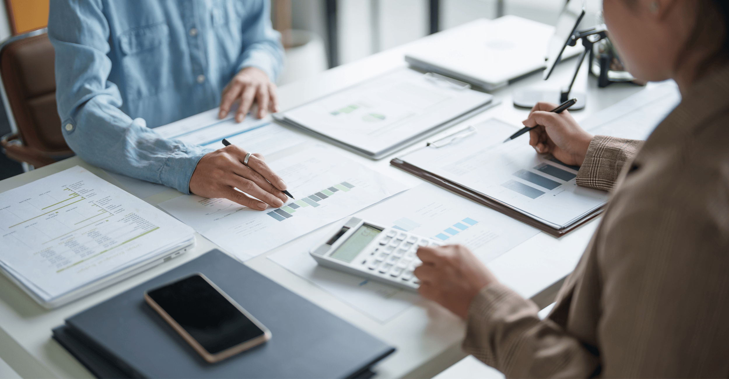 Close up of an organised set of documents showing bar charts and financial data, with two people working using a calculator and pens.