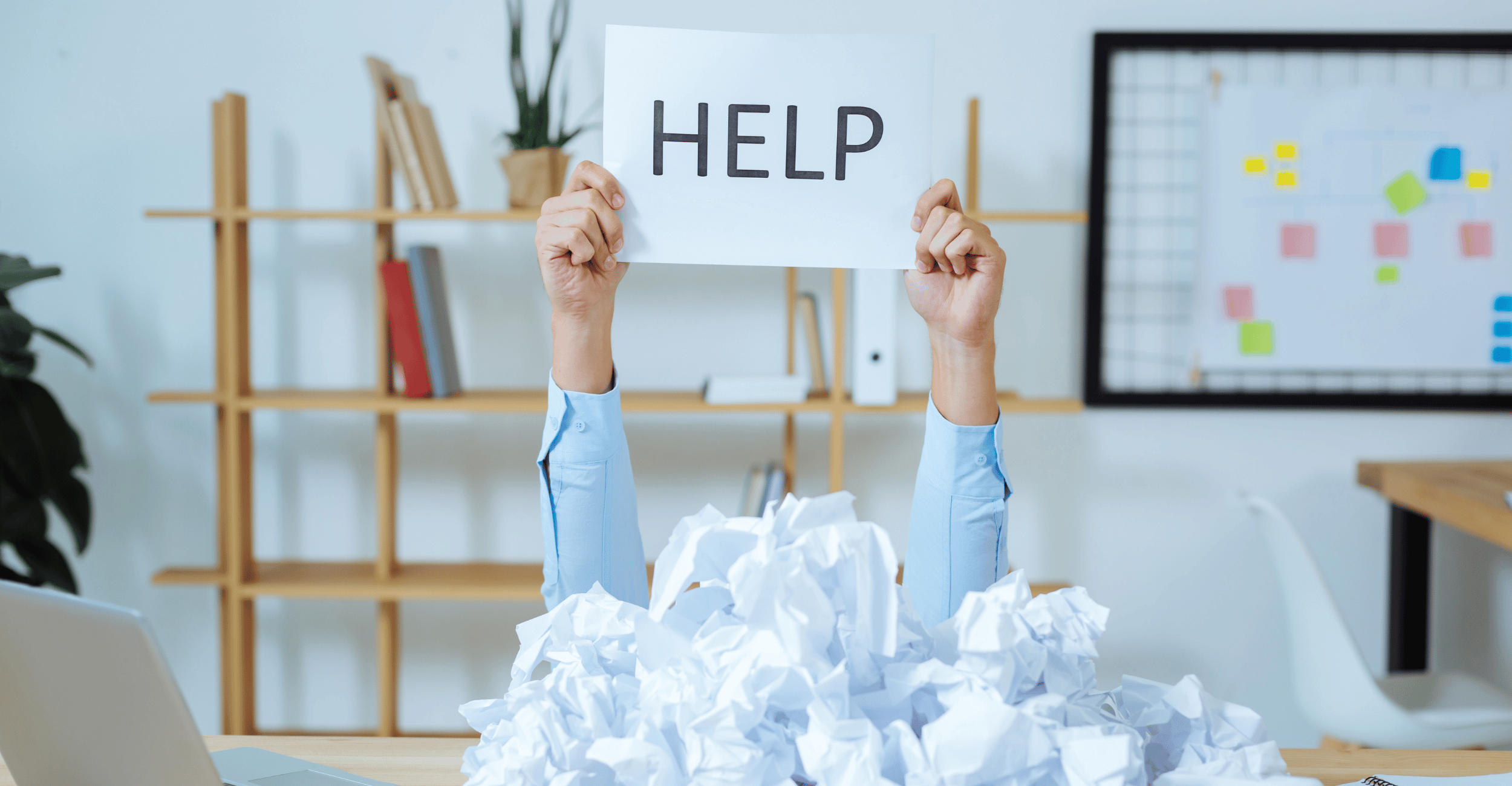 Two arms raised out of a pile of scrunched up paper, holding a sign saying Help. The person is in an office environment.