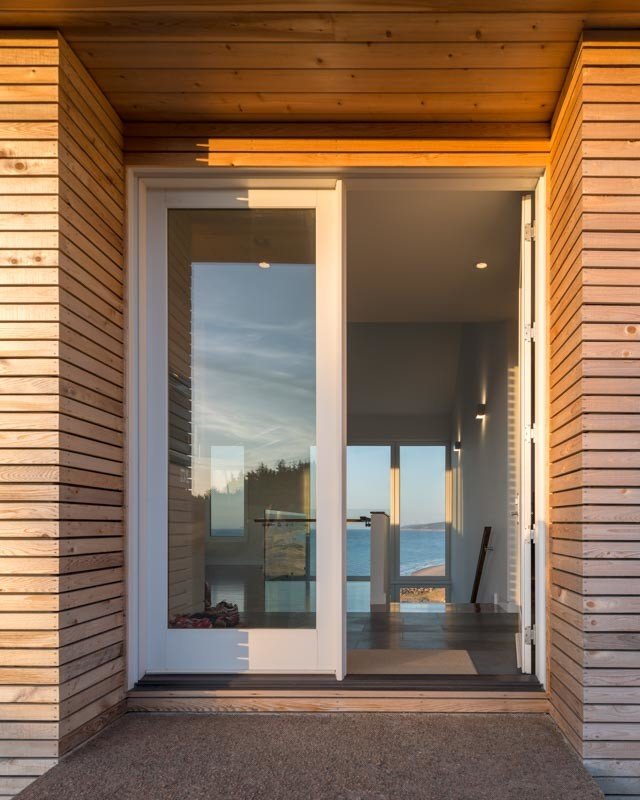 Modern wooden house entrance with open door, interior view of ocean through large windows.