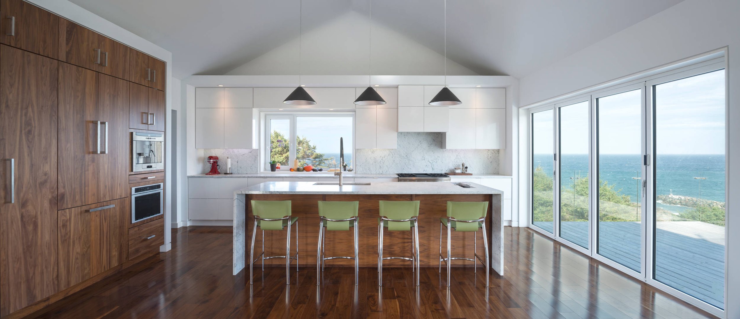 Modern kitchen with wooden cabinets, a white island with green chairs, large windows with ocean view, and pendant lights overhead.