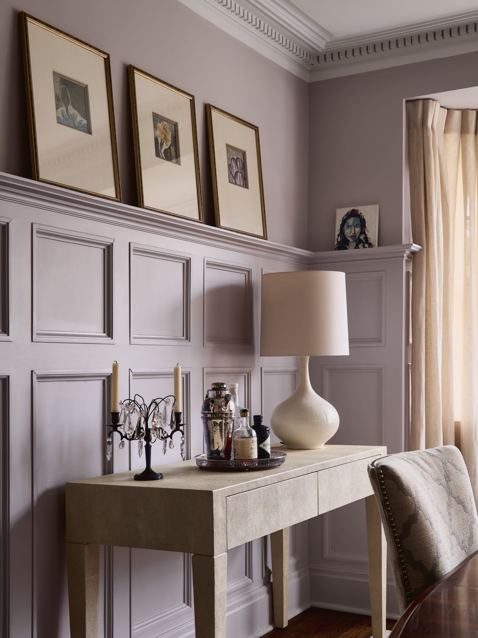 Elegant room with a beige console table against paneled walls, adorned with framed artwork. A decorative chandelier-style candle holder, a large white lamp, and a tray with bottles and a cocktail shaker sit on the table. Beige curtains cover large wi