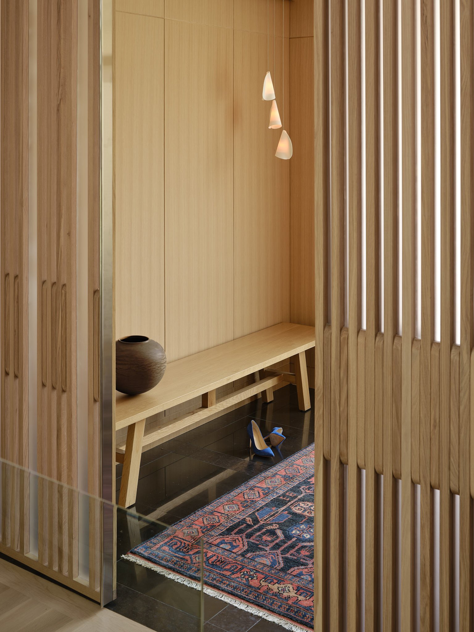 Minimalist wooden interior with bench, pendant lights, patterned rug, clay vase, and blue high-heeled shoes.