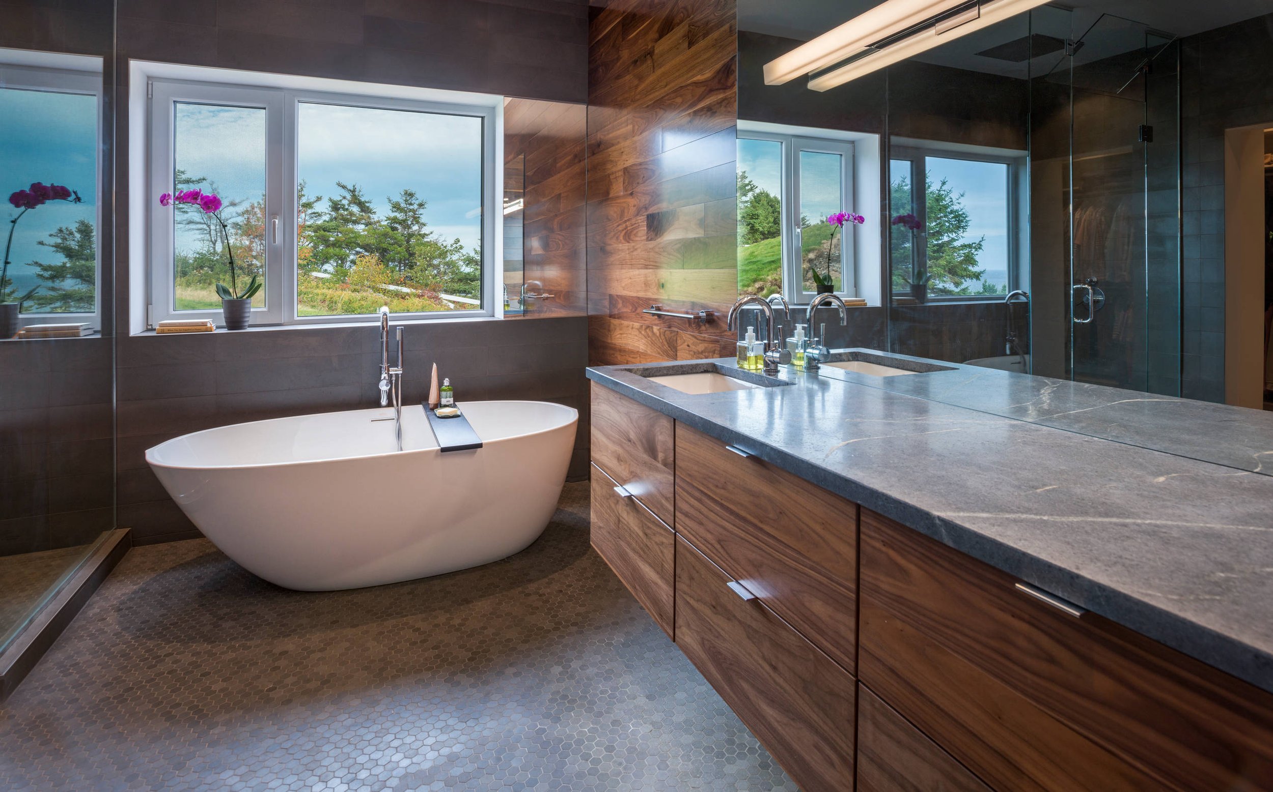 Modern bathroom with freestanding bathtub, dual sinks, wooden cabinets, large windows, and orchid plants.
