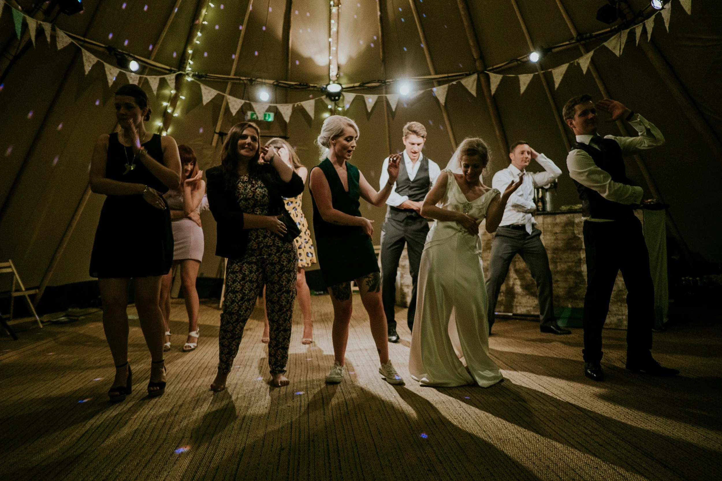 People dancing at a celebration or party inside a large tent with string lights and bunting decorations.