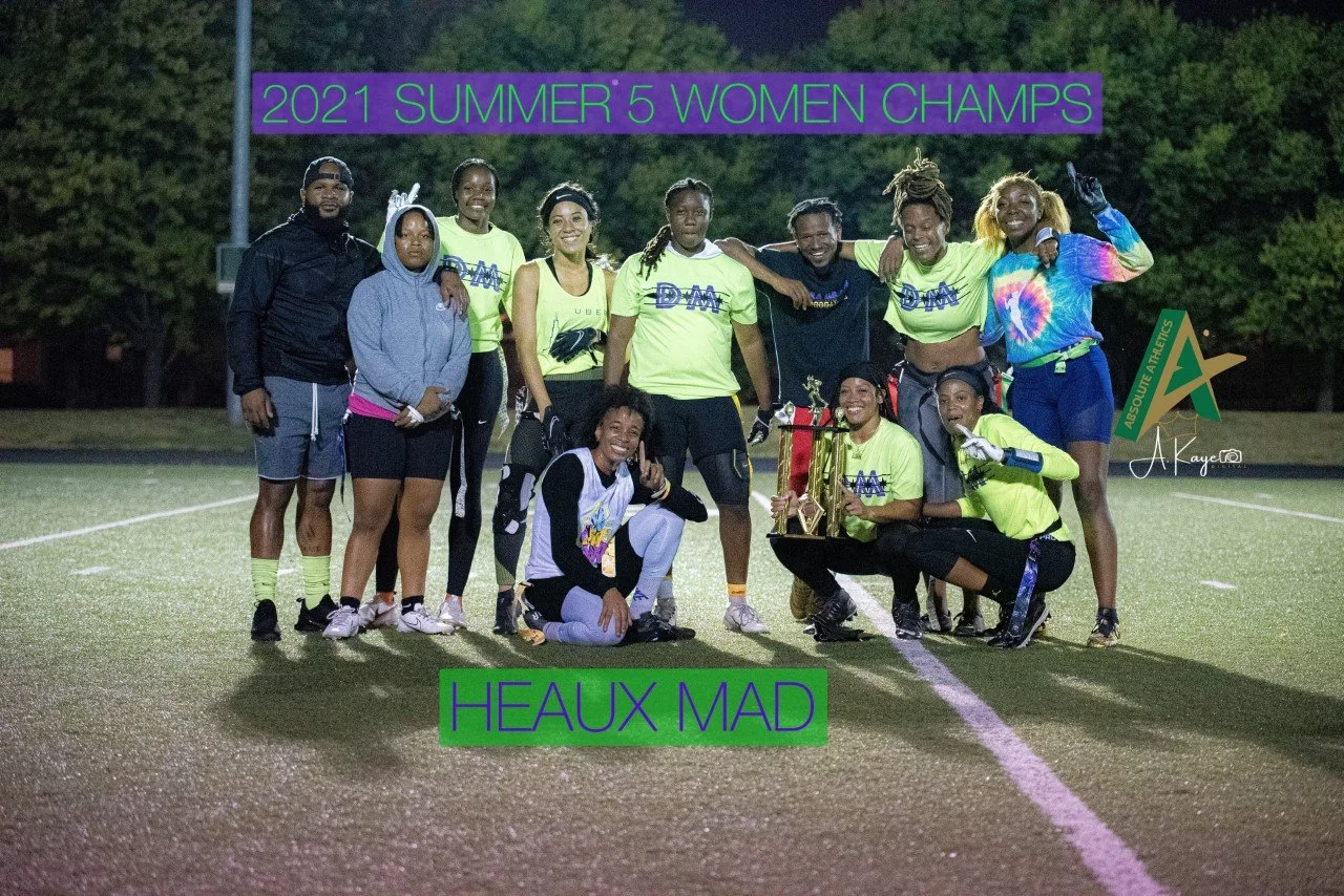 A group of women and men celebrating on a sports field at night, holding trophies and wearing matching bright yellow shirts that read 'DM.' The text on the image announces they are the 2021 Summer 5 Women Champs, and the bottom text reads 'Heaux Mad.'