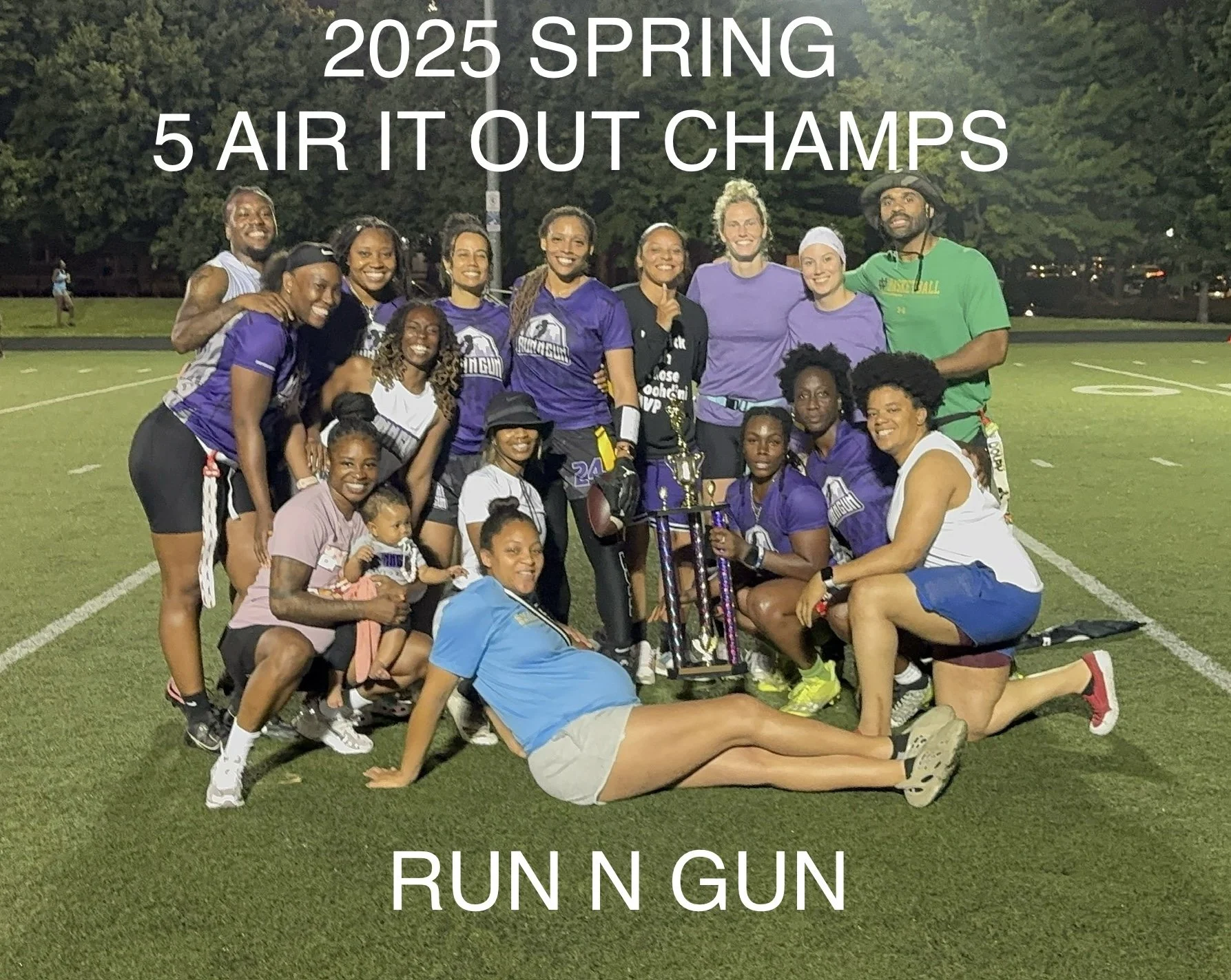 Group of women and girls on a football field celebrating with a trophy after a spring 2025 Air It Out Champs race event, some wearing athletic gear, smiling, and holding medals, with a background of trees and a night sky.