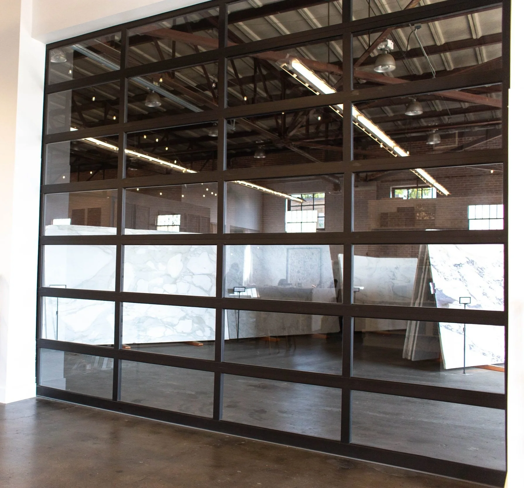 Interior of an art gallery or showroom with large display panels showing marble patterns. The area has brick walls, high ceilings with exposed beams, and industrial lighting. Seen through a glass window with black framing.