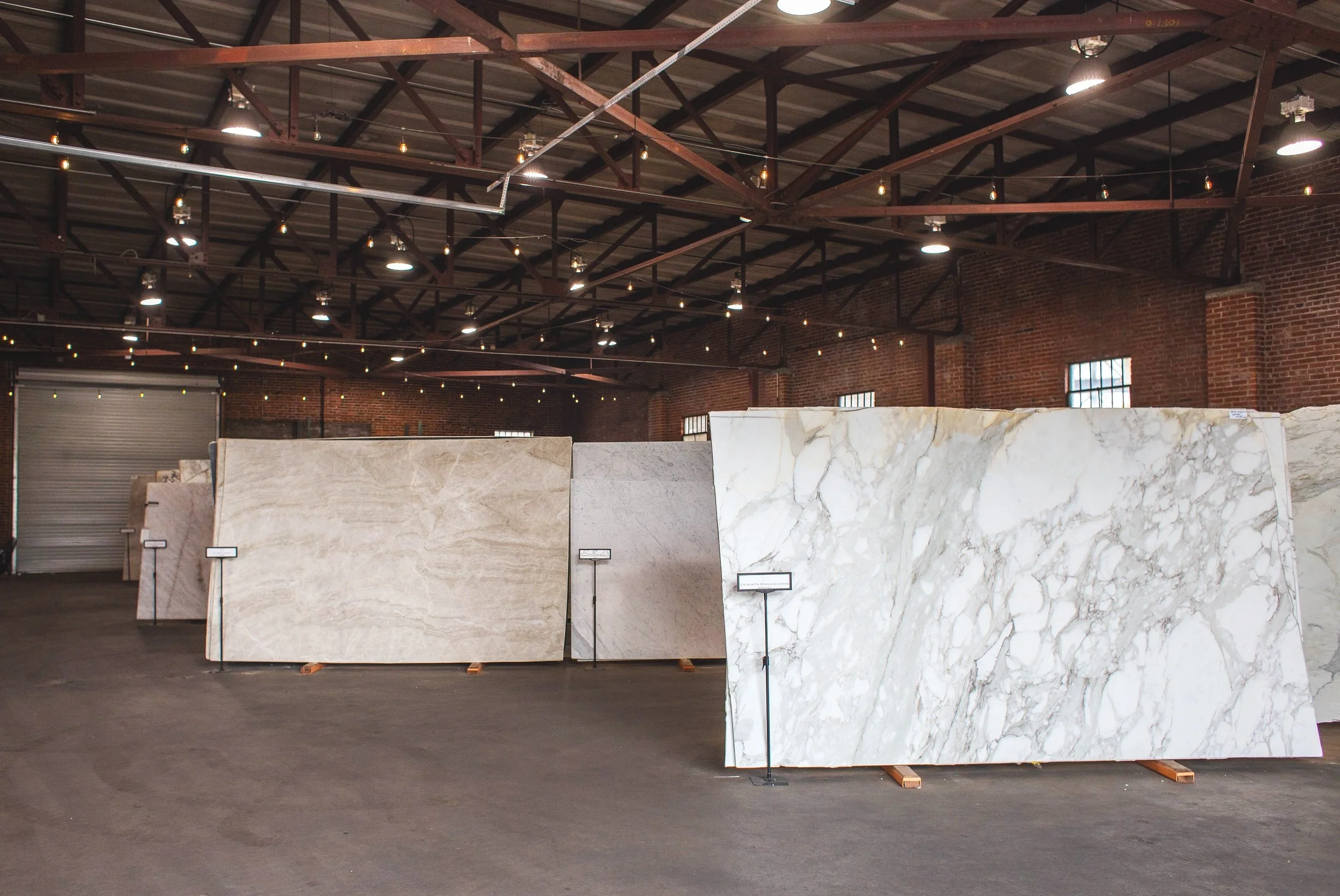 Large slabs of marble displayed in a warehouse with brick walls and string lights overhead.