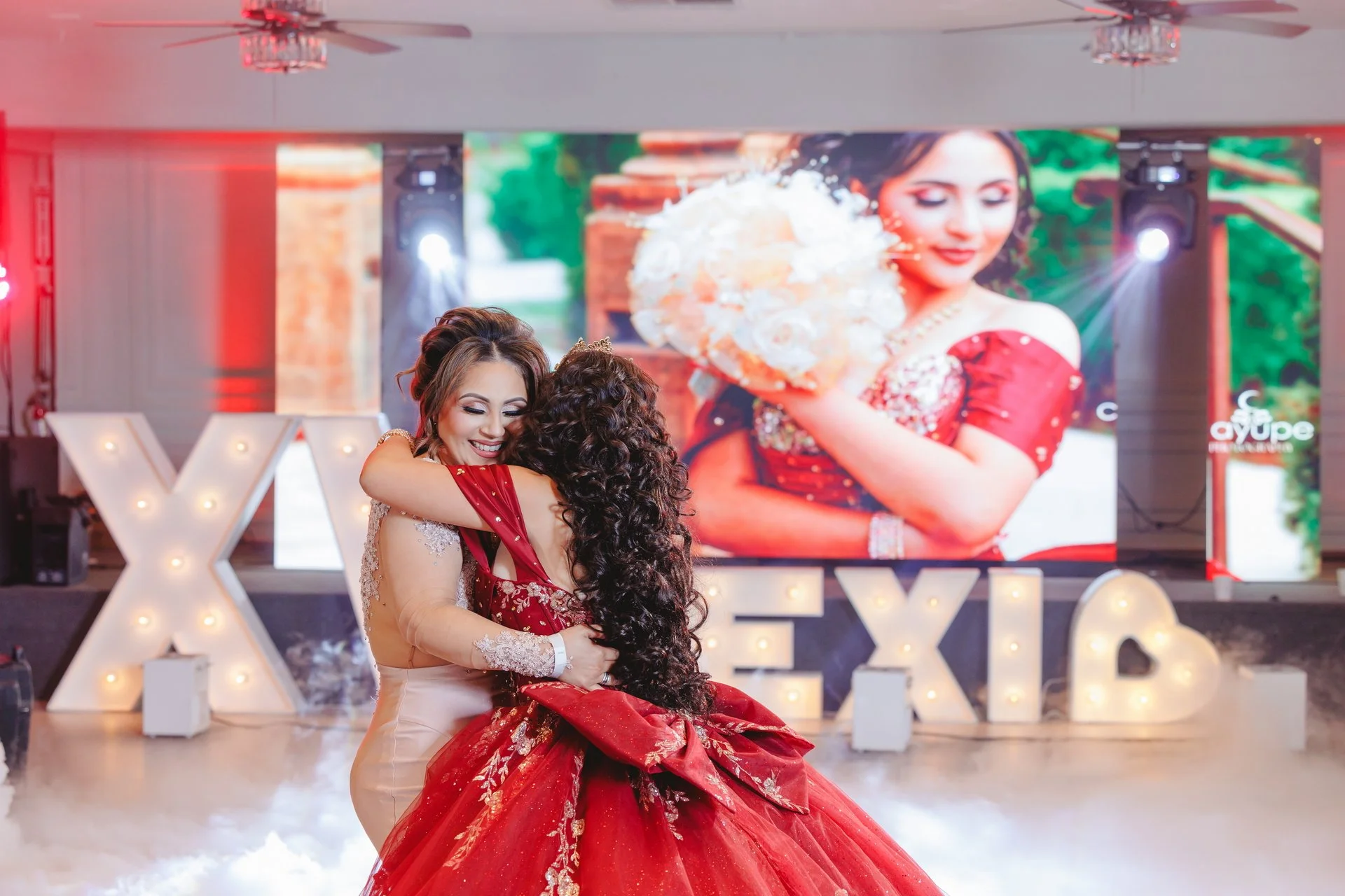 Quinceneara in elegant red gown embracing woman in front of beautiful decorations at event venue