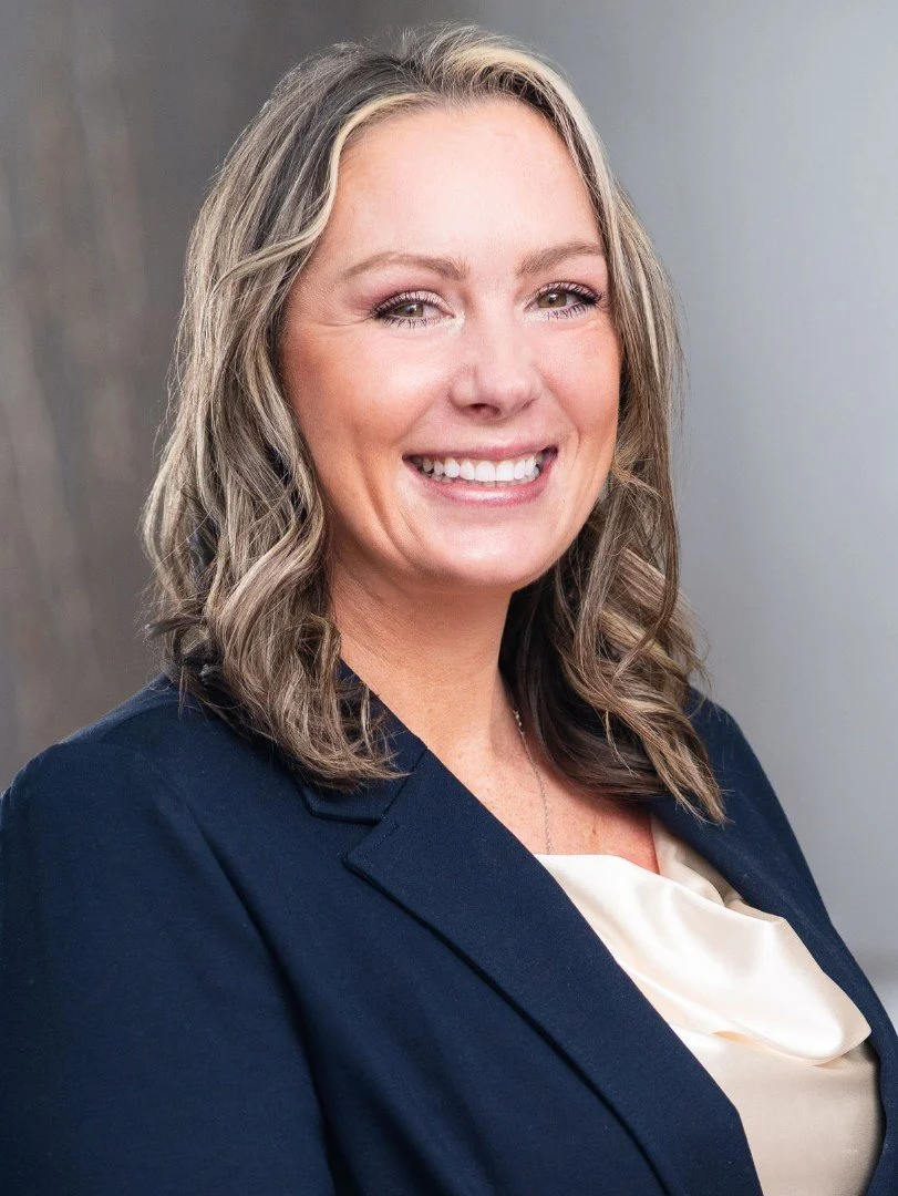 A woman with shoulder-length wavy blonde hair smiling, wearing a navy blazer and a light-colored blouse against a neutral background.