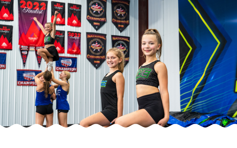 Four young cheerleaders practicing stunts inside a gym, with banners on the wall celebrating cheerleading championships from 2021 and 2022, and a large blue wall with yellow and black design elements.