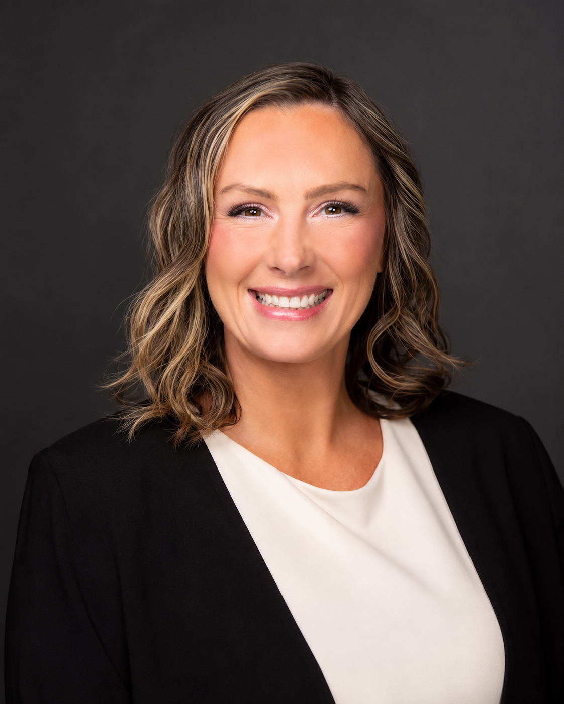 A woman with long, wavy, light brown hair, blue eyes, and fair skin, smiling, wearing a black blazer over a teal patterned blouse and a necklace with a feather pendant, against a gray background.