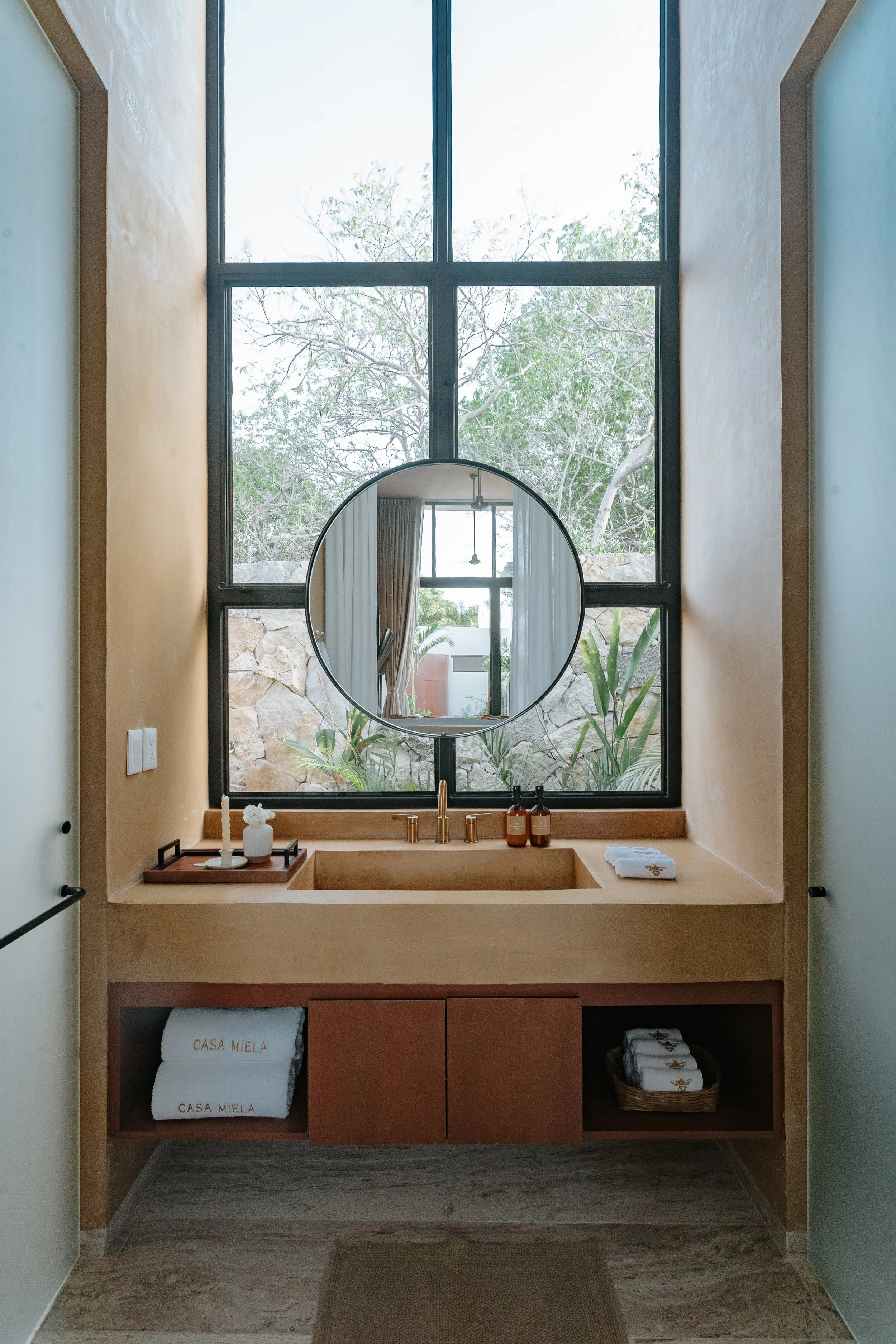 Bathroom vanity with a large window, round mirror, sink, and toiletries, overlooking greenery outside.