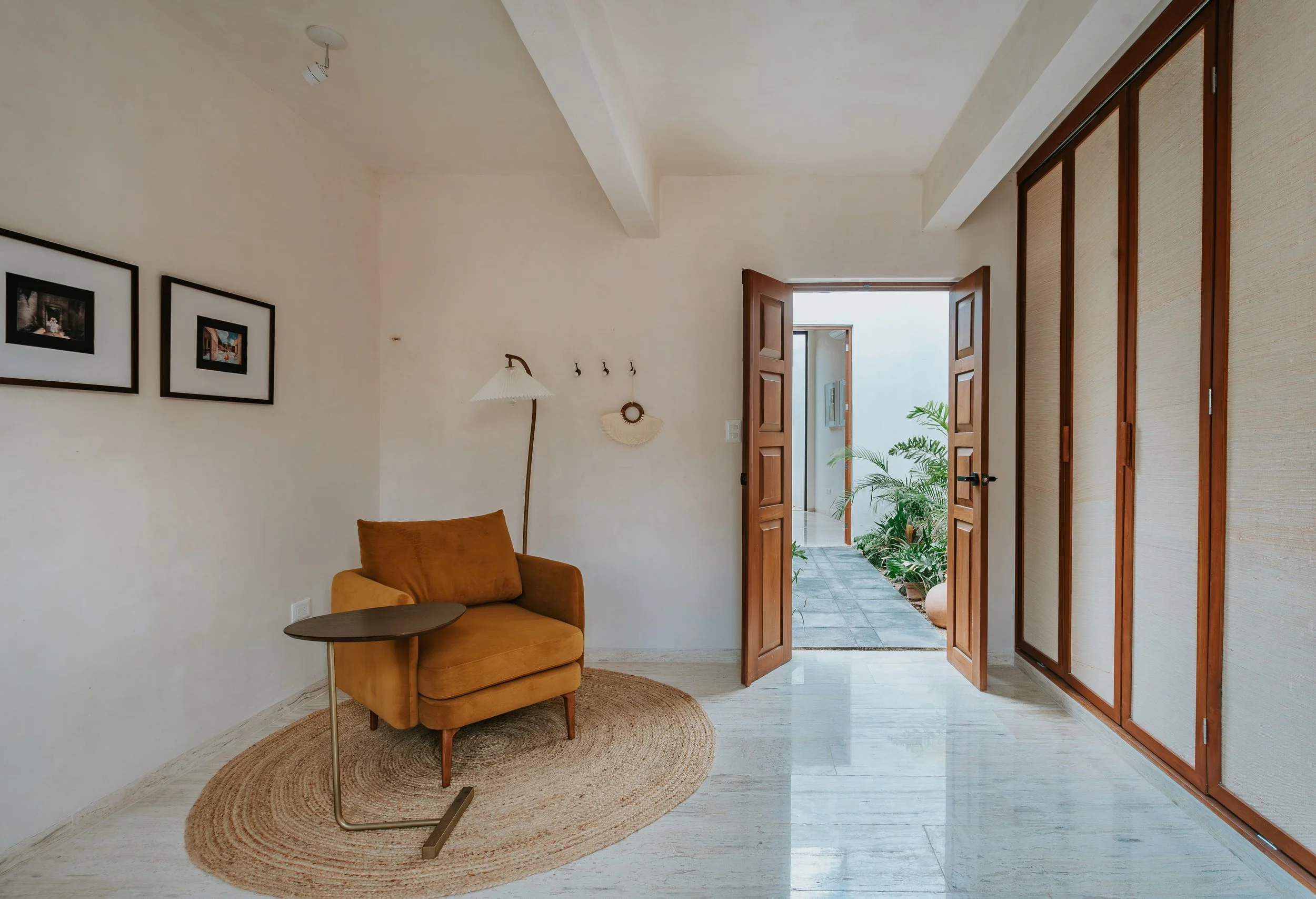 A cozy living room with a mustard yellow armchair on a circular jute rug, a tall floor lamp with a white shade, and framed photographs on the wall. Double wooden doors open to a small outdoor patio with potted plants.