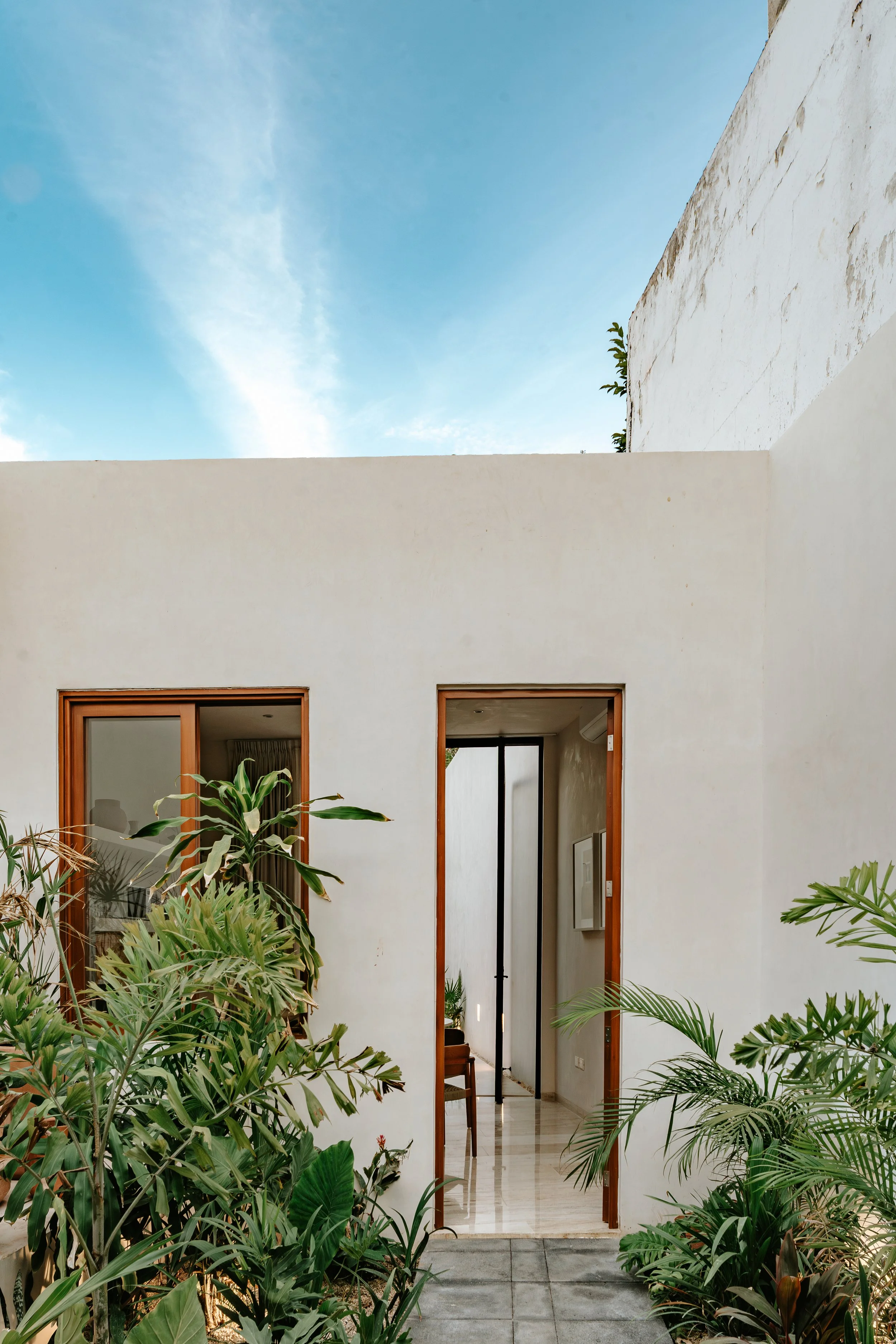 Architectural view of a modern building entrance with white walls, open doorways, and lush green plants outside under a blue sky. colonial house merida, restoration project
