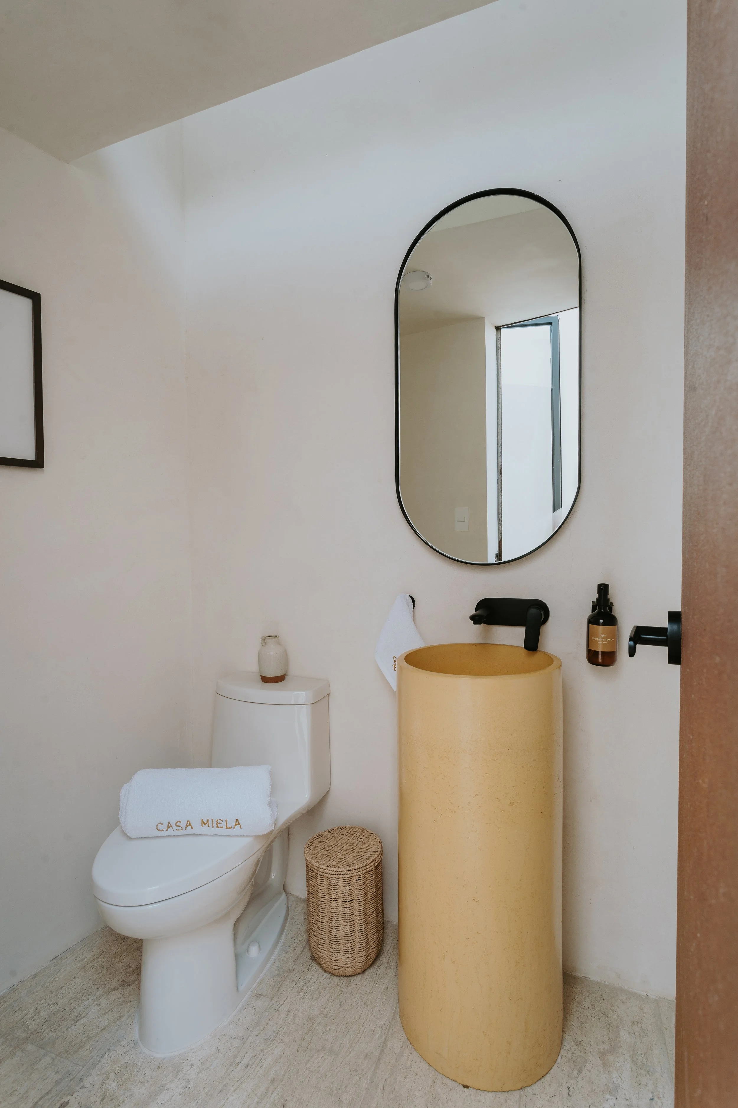 Modern bathroom featuring a white toilet with a towel labeled 'Casa Miela', a beige cylindrical sink, a large mirror, a black faucet, a small wicker waste basket, and bottles on the wall.