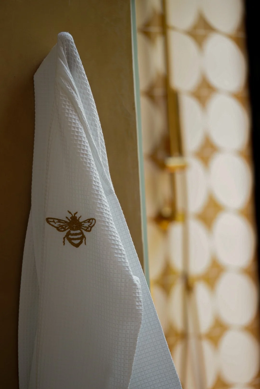 A white textured bathrobe hanging on a hook on a beige wall, with a golden honey bee embroidery on it. Part of a room with warm tones and a decorative wall panel in the background.