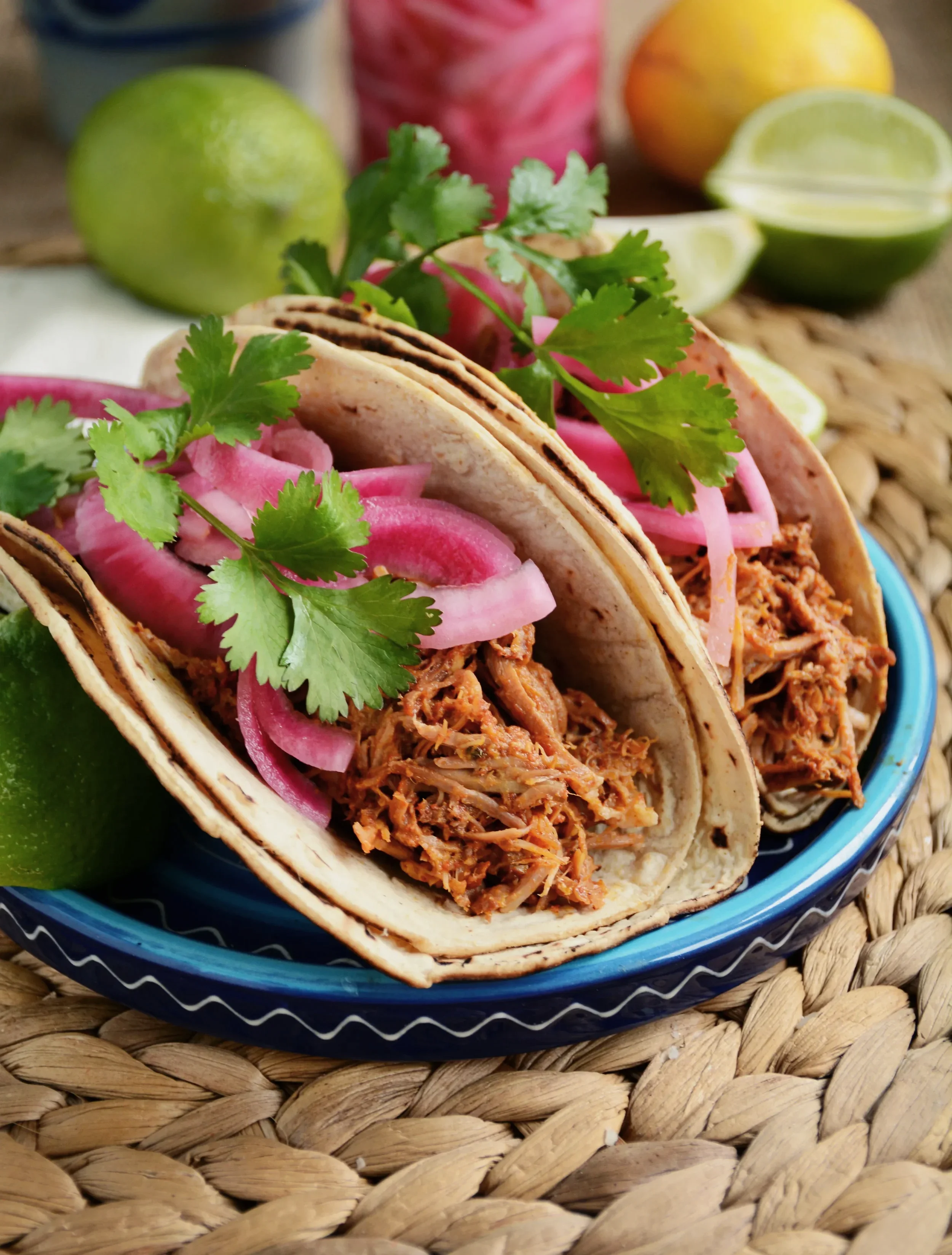 Two tacos filled with shredded meat, pickled onions, and cilantro in a blue bowl on a woven mat, with limes and other fruits in the background.