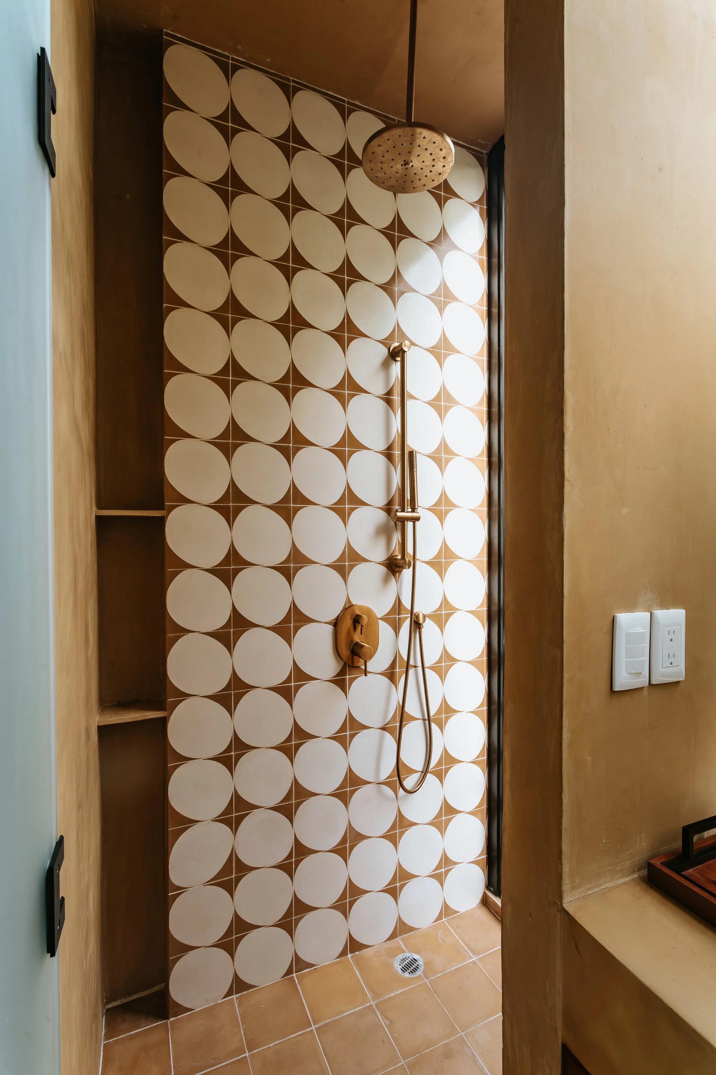 Shower area with a rainfall shower head, a handheld shower, brown and white patterned tiles, built-in shelves, and a floor drain.