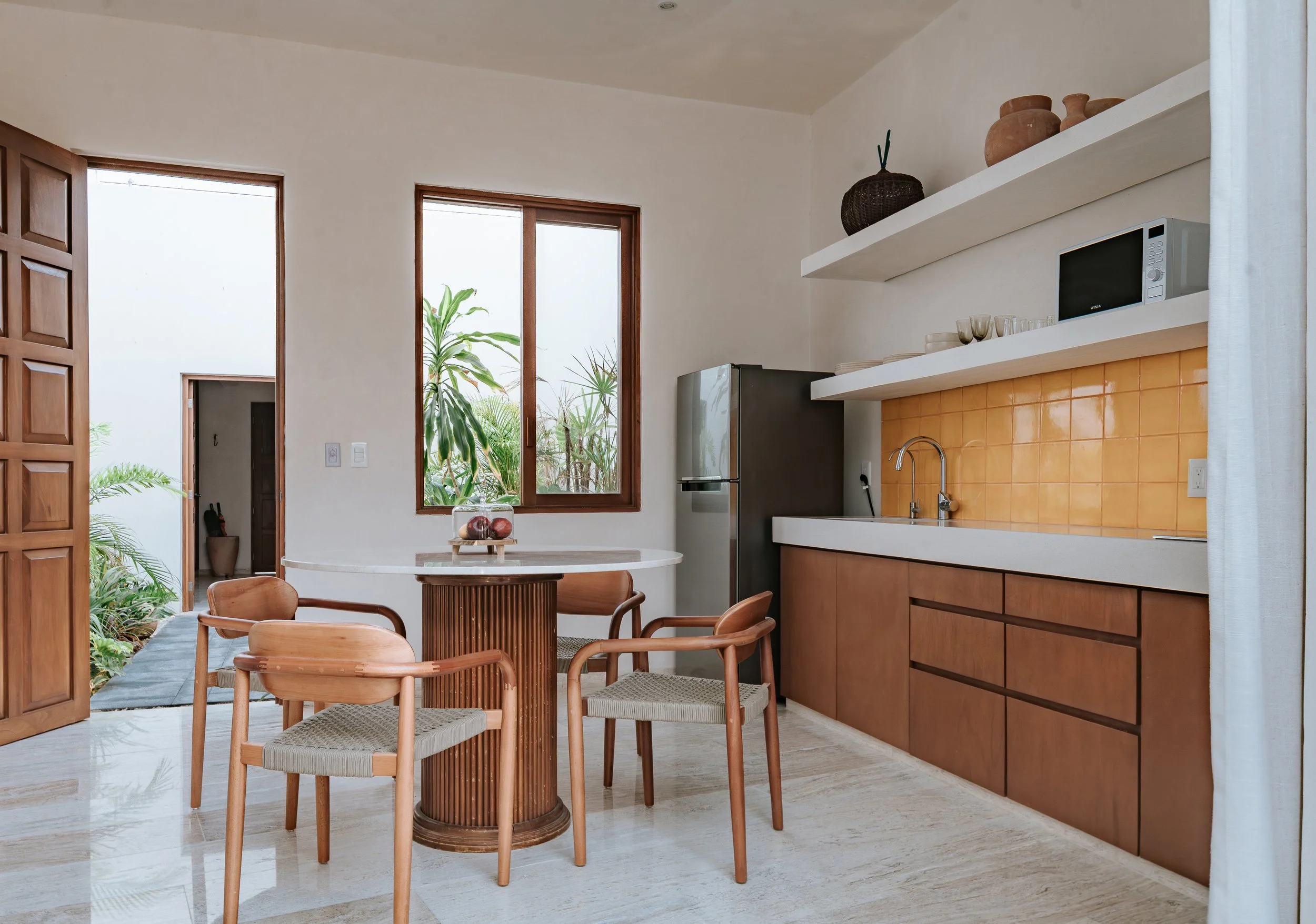 A kitchen with a small round dining table and four wooden chairs, a refrigerator, open shelves with bowls and pottery, a microwave, a yellow tiled backsplash, and a large window with plants outside.