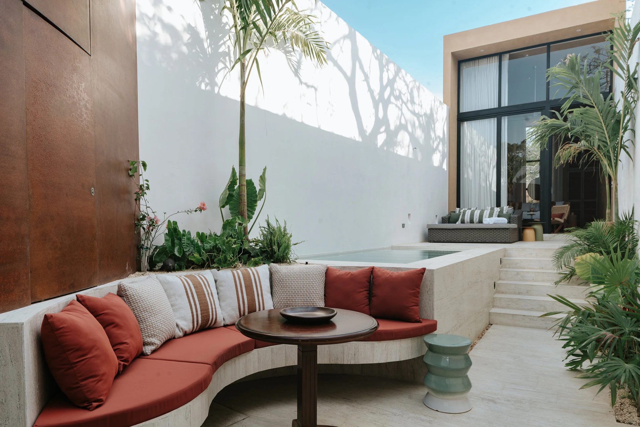 Outdoor patio area with built-in curved seating with red cushions, a small round wooden table, potted plants, and a swimming pool, illuminated by natural sunlight with shadows of trees on the white wall.