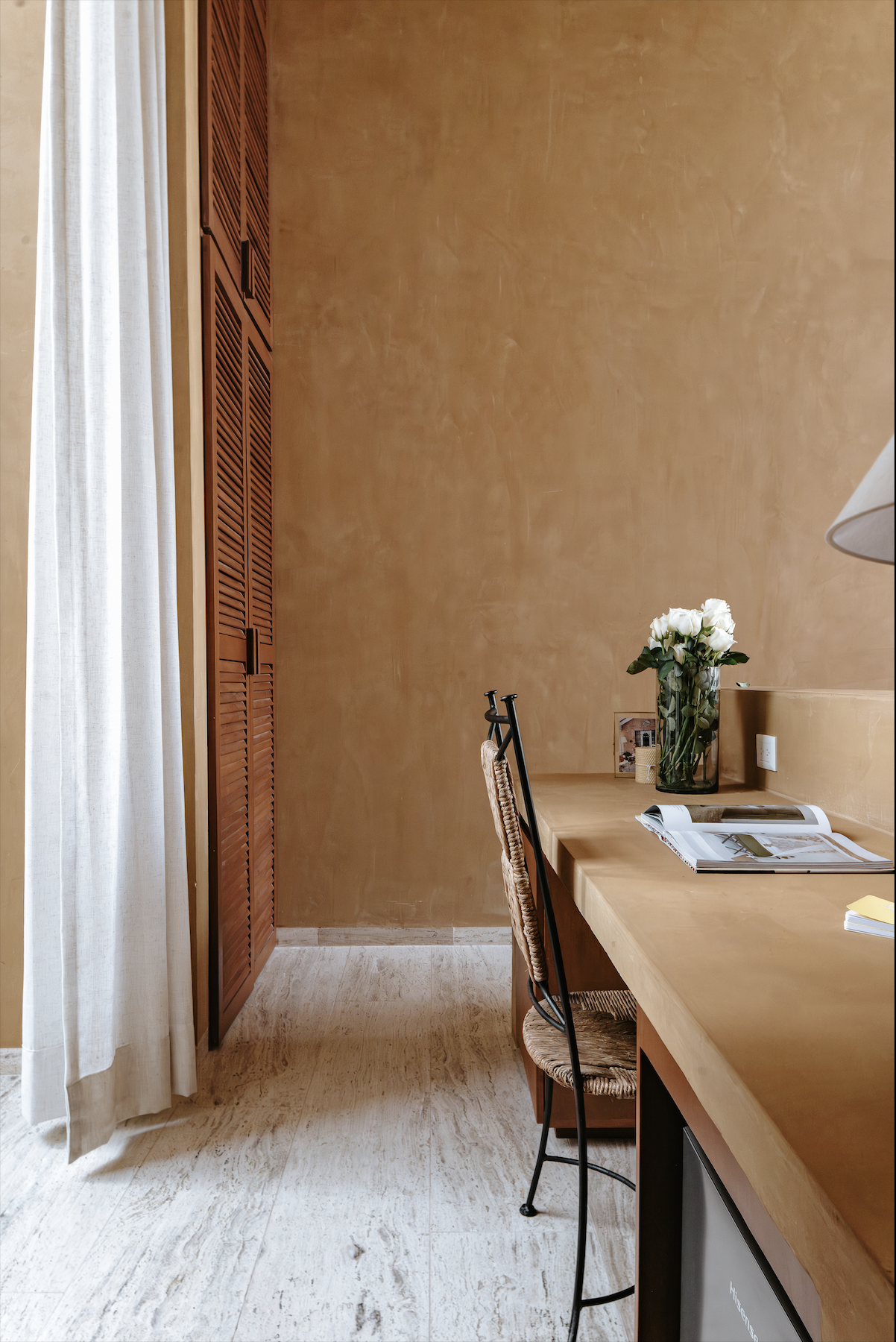 A corner of a room with a wooden desk, a black chair with a woven seat, a vase of white roses, some open magazines, a small picture, and a wall with a textured beige finish.