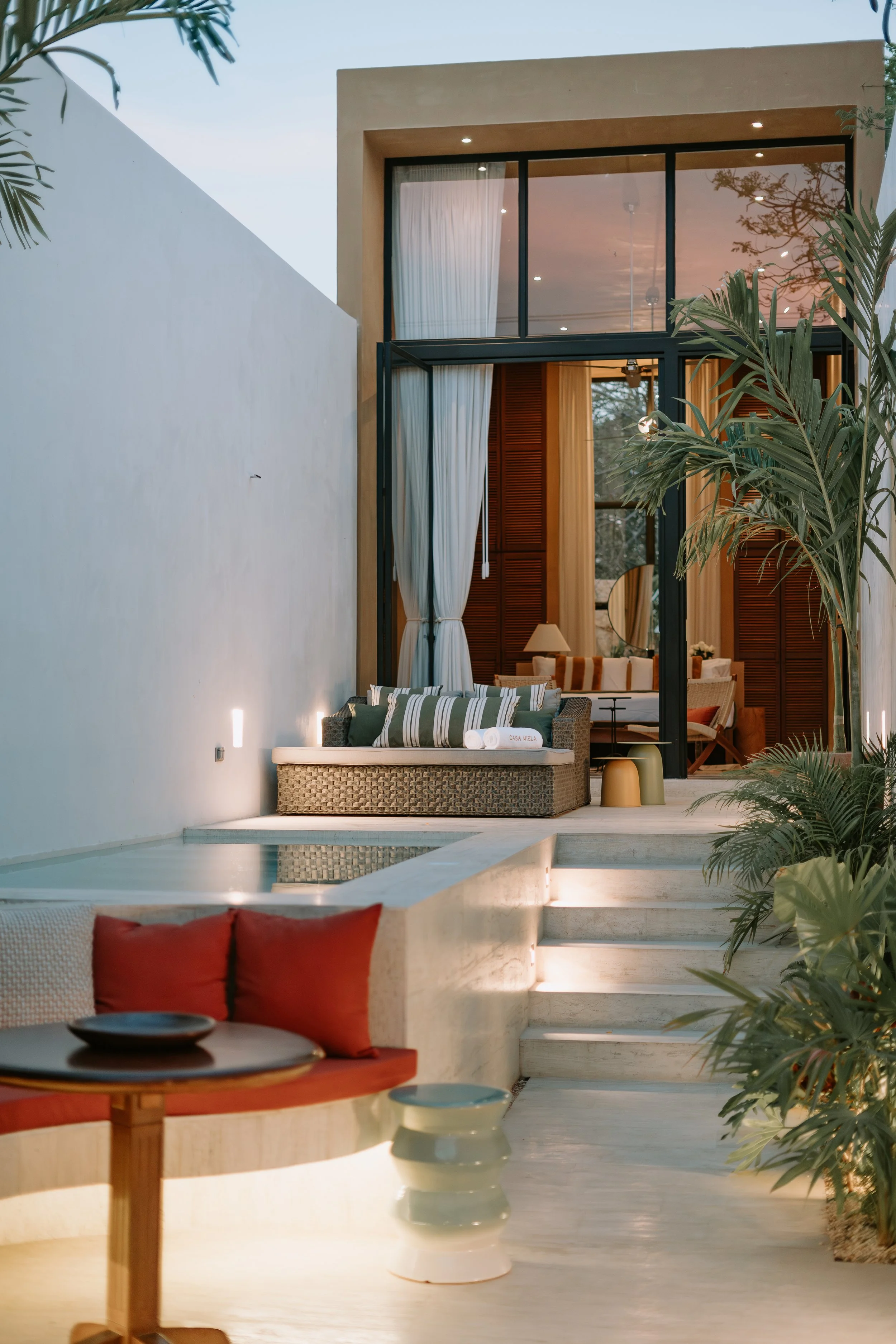 Modern backyard area with a small pool, outdoor seating with cushions, a table, and lush green plants. The house interior is visible through large glass doors, showing a cozy living room with wooden accents and curtains.