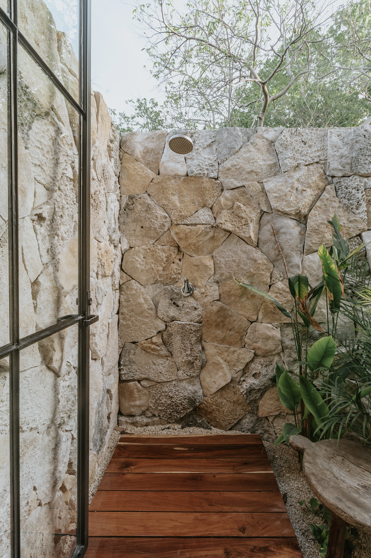 Outdoor stone shower with a wooden floor, a rain showerhead, a small side table made of wood, and green plants on the right, with trees visible above the stone wall.