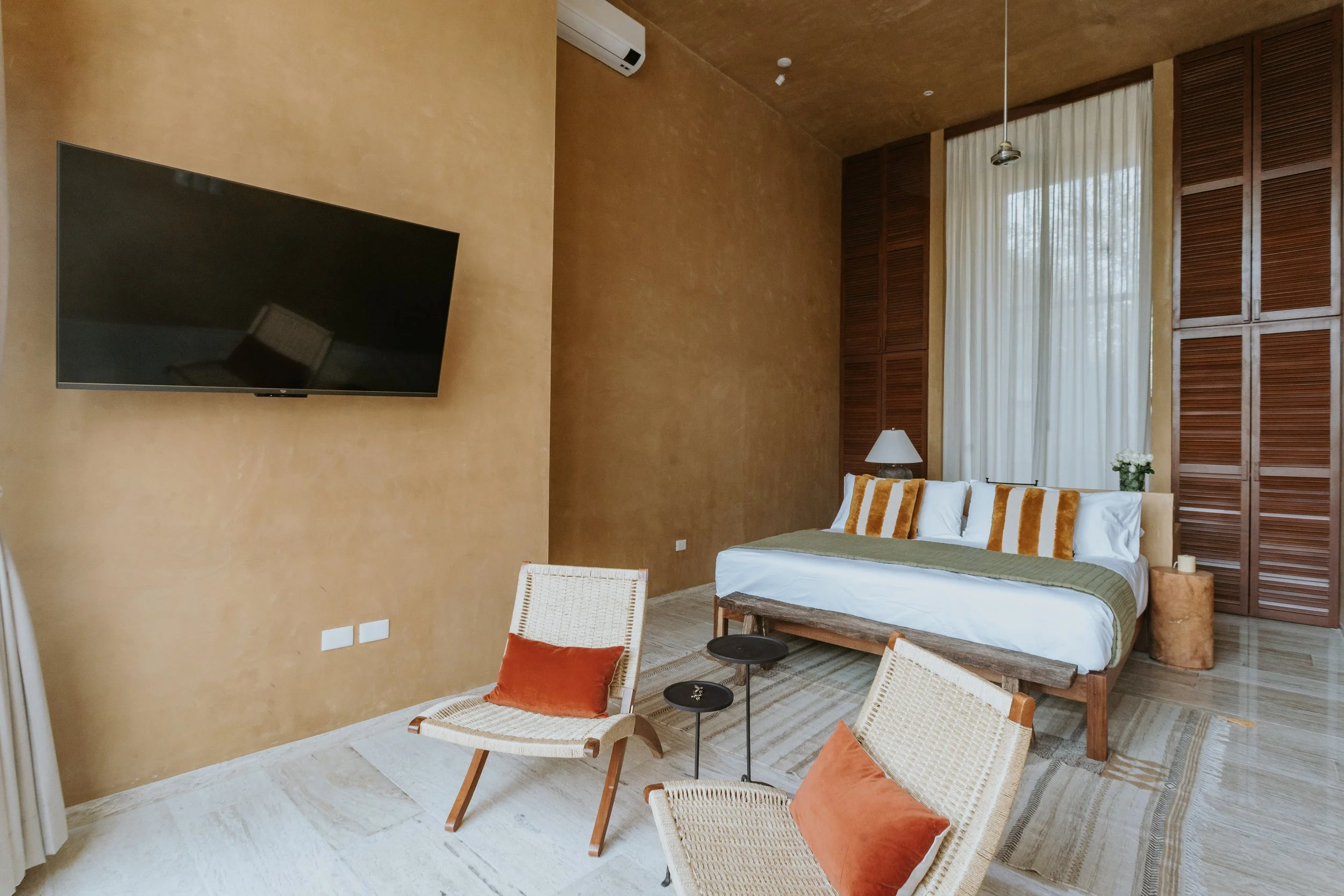 A spacious hotel bedroom with a large bed covered in white sheets, green blanket, and striped pillows. There are two woven chairs with orange pillows and small black side tables. A flat-screen TV is mounted on a beige wall, and there is a large windo