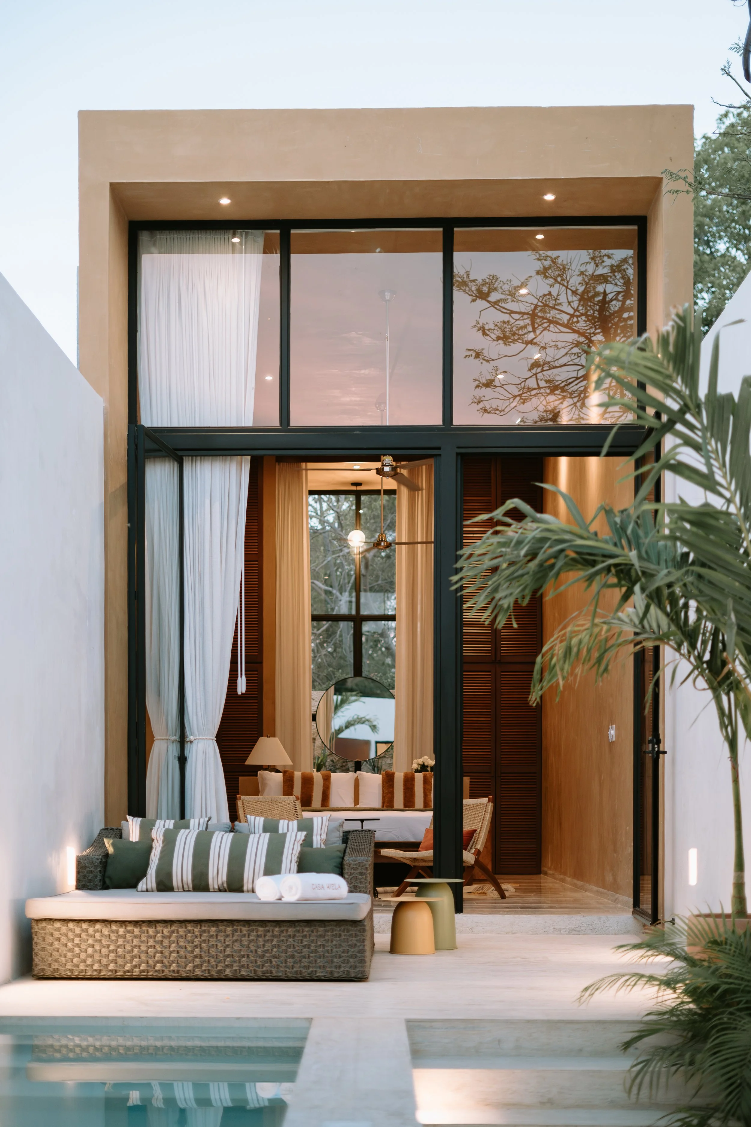 Modern house with large glass windows and sliding doors, outdoor seating area with a woven sofa and throw pillows, a pool in the foreground, and lush greenery around.